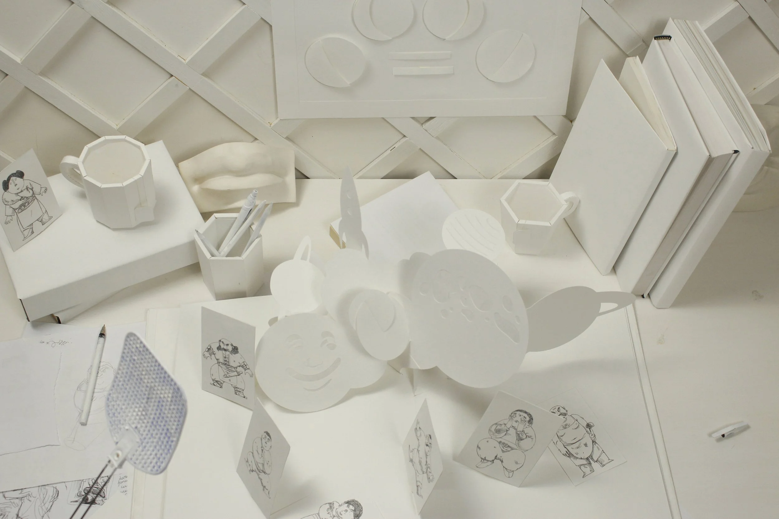 White desk with various paper cutouts, drawings, books, and office supplies arranged on it, including a paper honeycomb, a mouth sculpture, and illustrated cards.