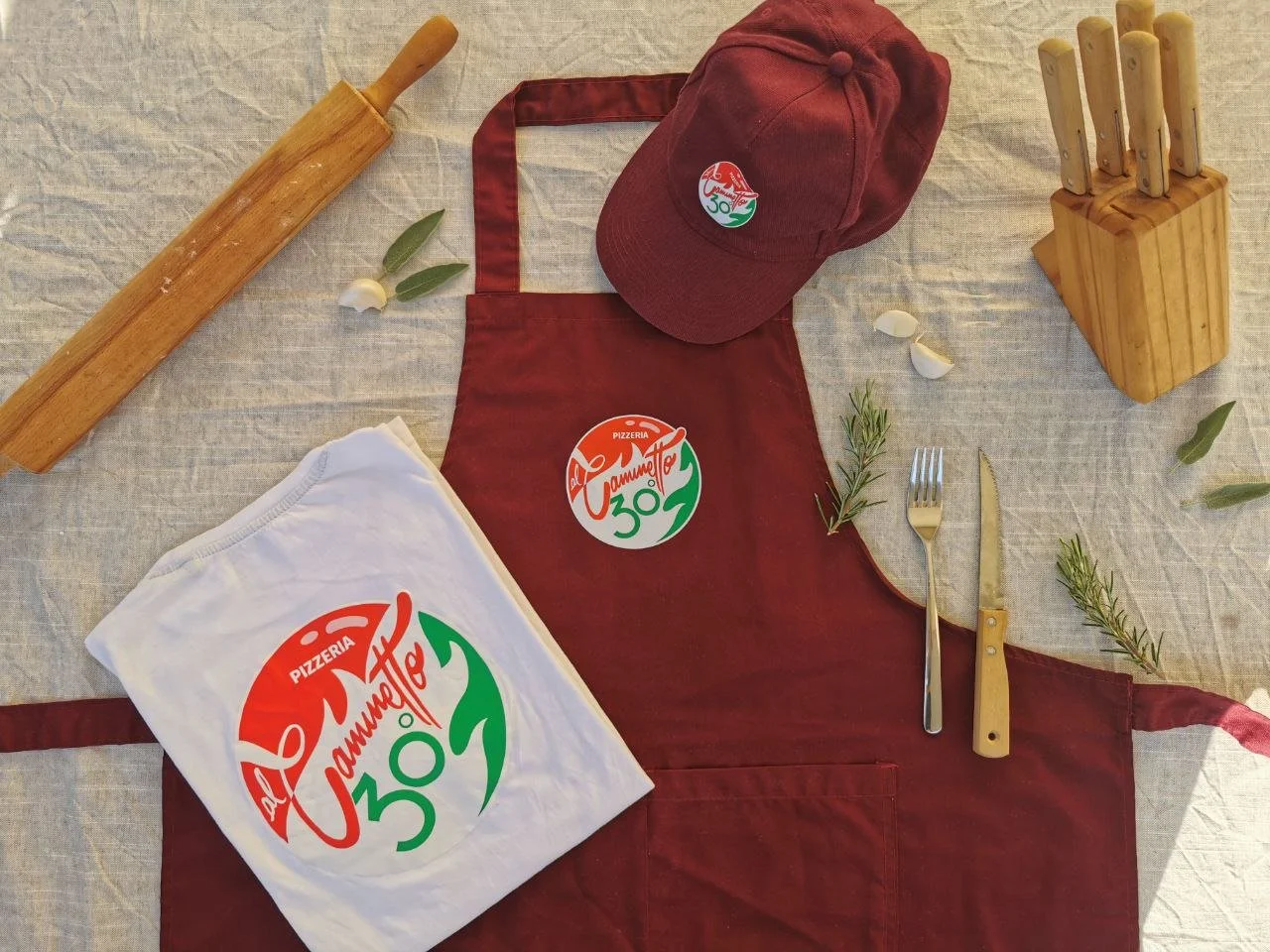 Tovaglia con grembiule, cappello, coltelli, forchetta, pasta rolling pin, e foglie di rosmarino e alloro su una superficie