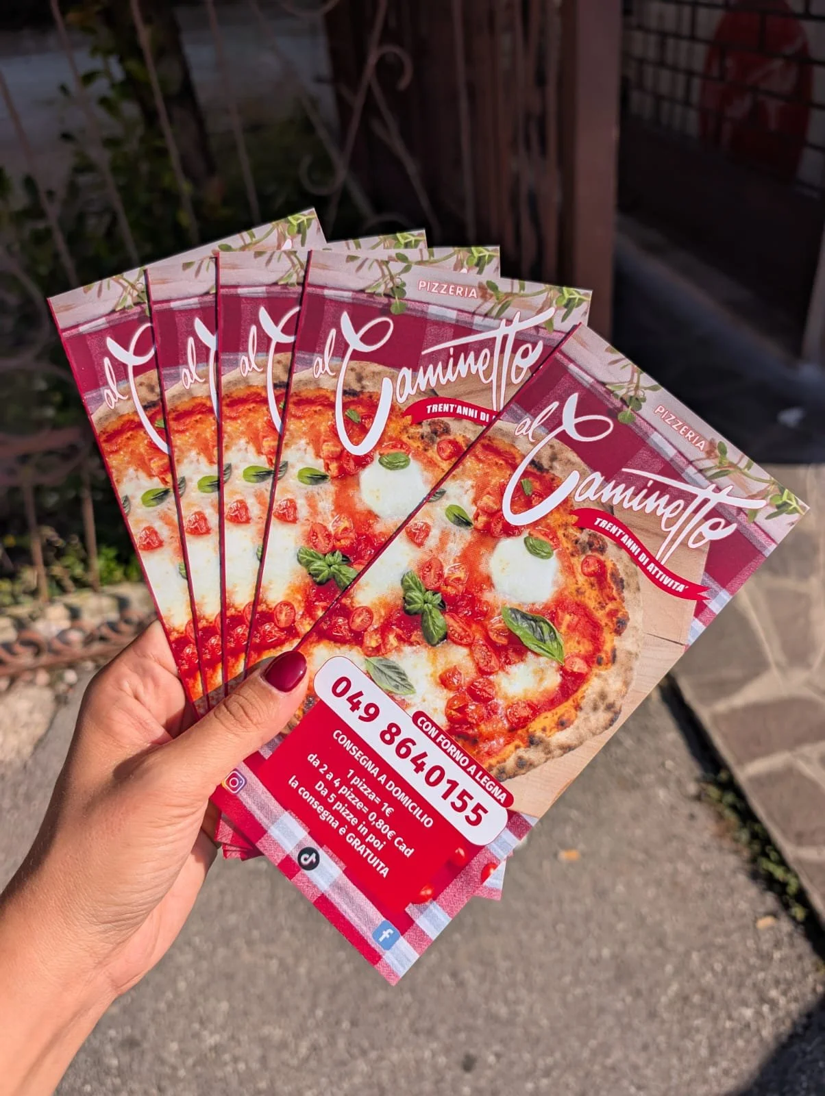 Mano che tiene cinque volantini pubblicitari di una pizzeria con immagine di una pizza margherita, all'aperto.