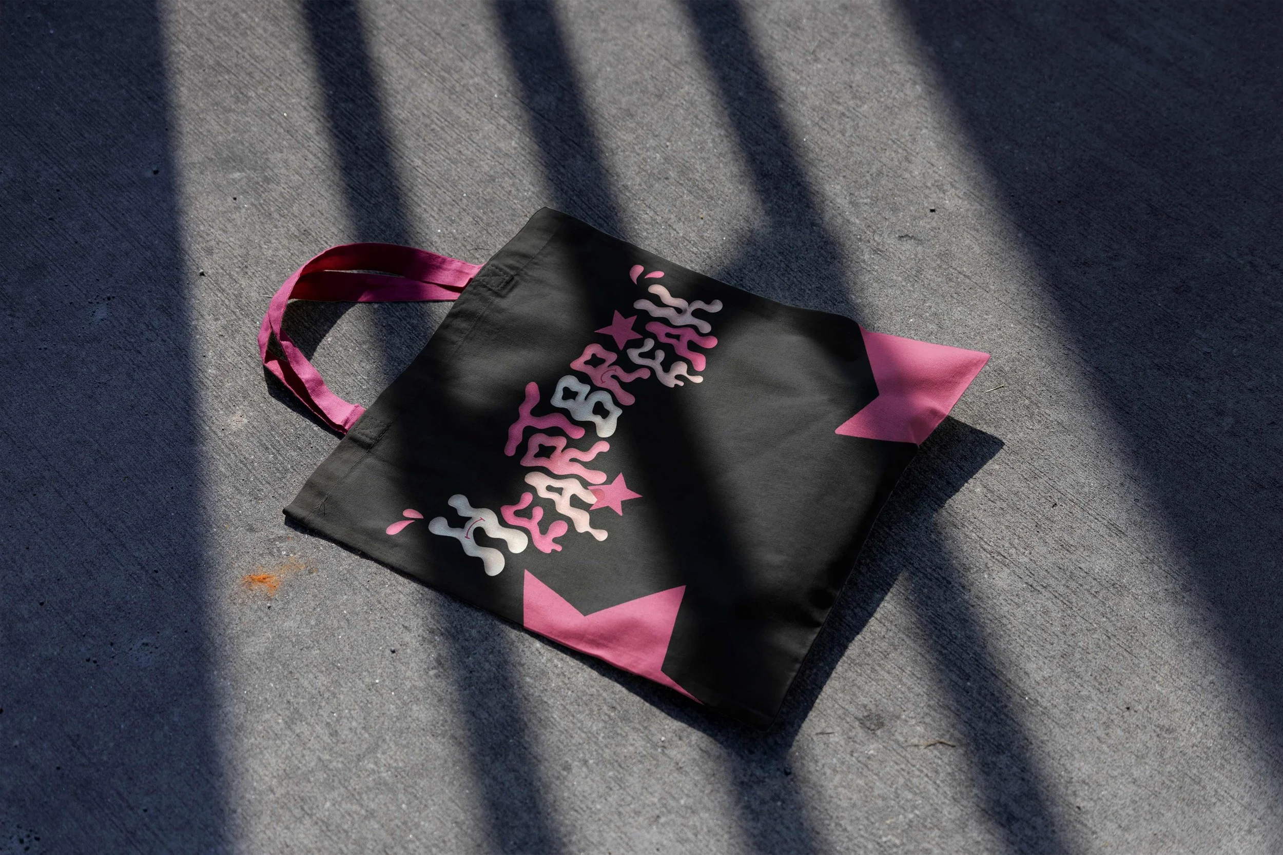Borsa di stoffa nera con dettagli rosa e scritte colorate, appoggiata su un marciapiede con ombre lunghe e diagonali.