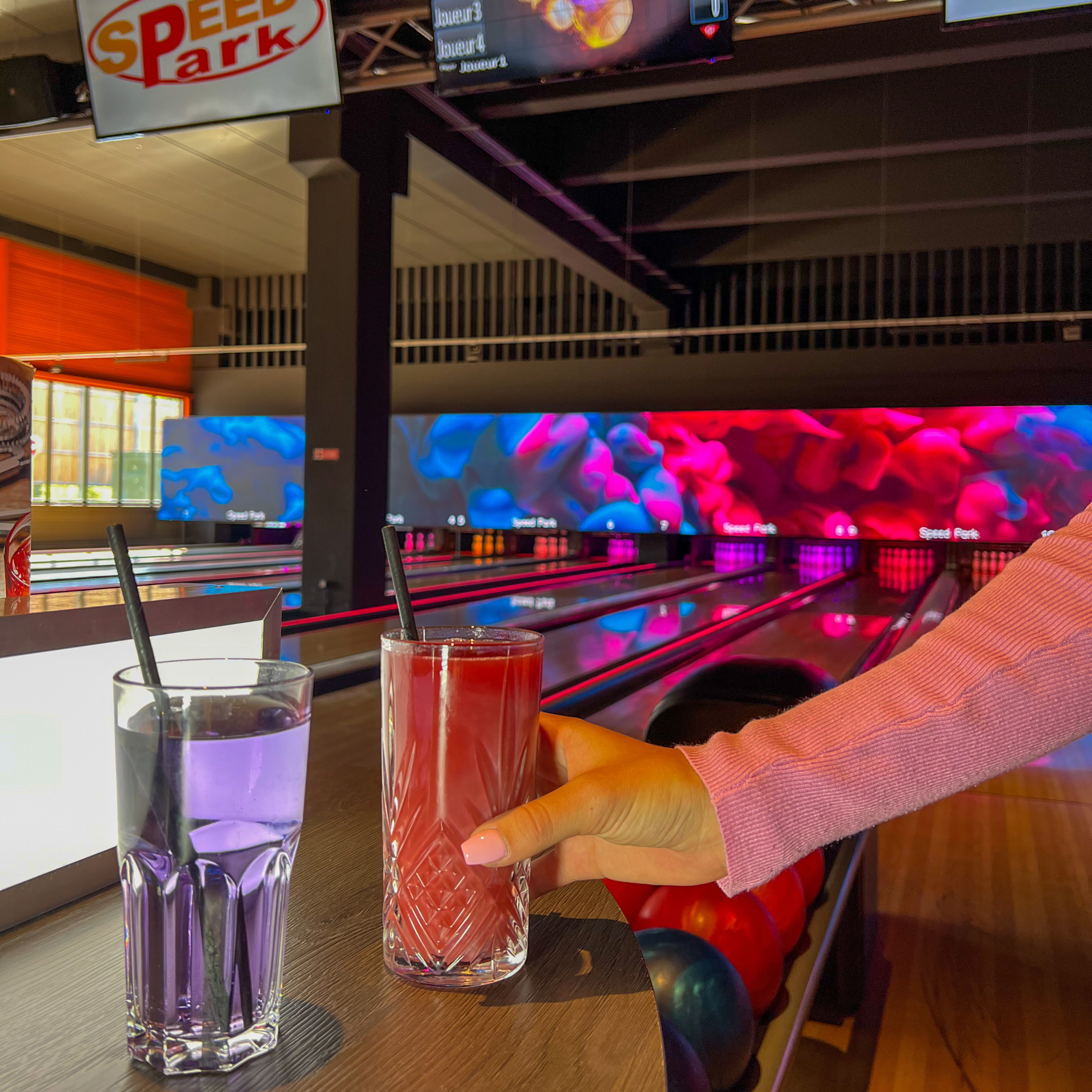 Une personne sert deux boissons colorées dans un bowling. Le fond montre des écrans de bowling avec des lumières vives et une piste de bowling avec des boules. Il y a un panneau 'Speed Park' en haut à gauche.