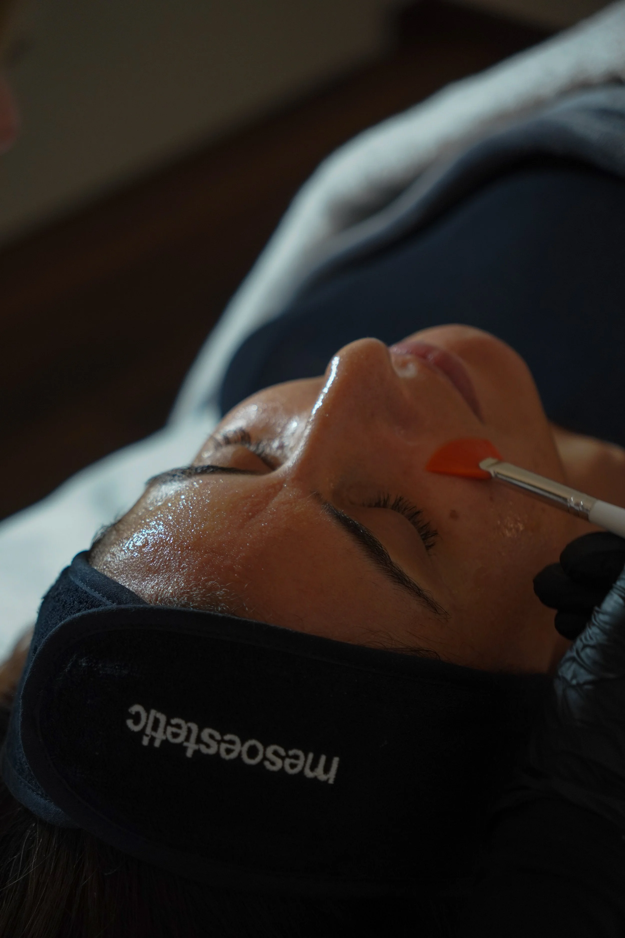 Une femme reçoit un soin du visage, allongée avec un bonnet de sommeil et un masque praticien appliquant un produit sur son nez.