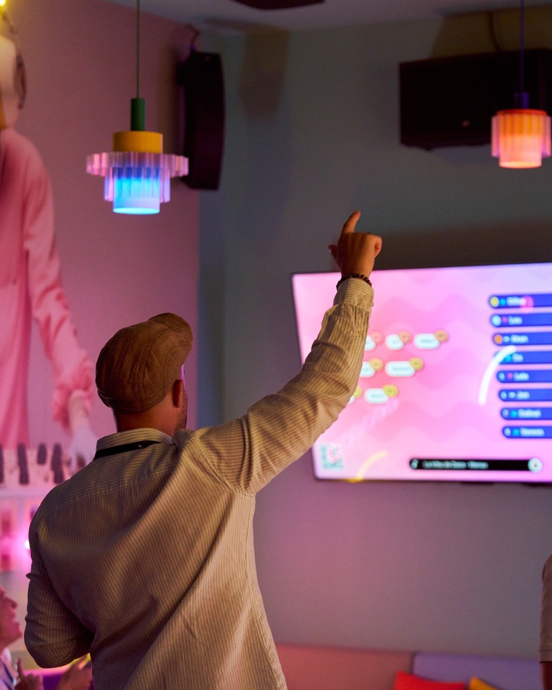 Un homme avec un chapeau beige et veste rayée, levant la main vers un écran numérique dans une salle avec un éclairage tamisé et des luminaires colorés suspendus au plafond.
