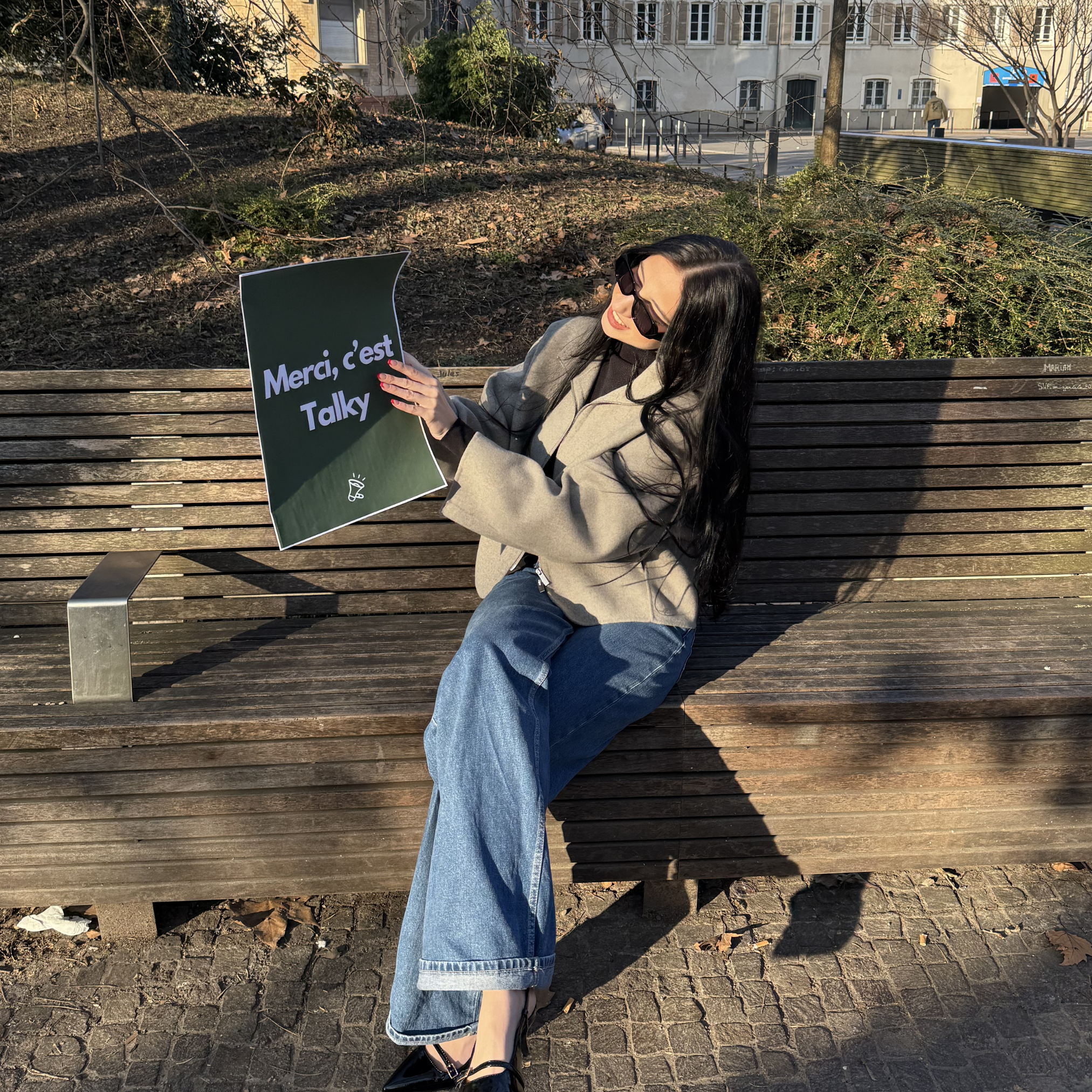Une femme assise sur un banc en bois, portant des lunettes de soleil, une veste beige, un pull noir et un jean large. Elle tient une affiche avec le texte 'Merci, c'est Talky'.