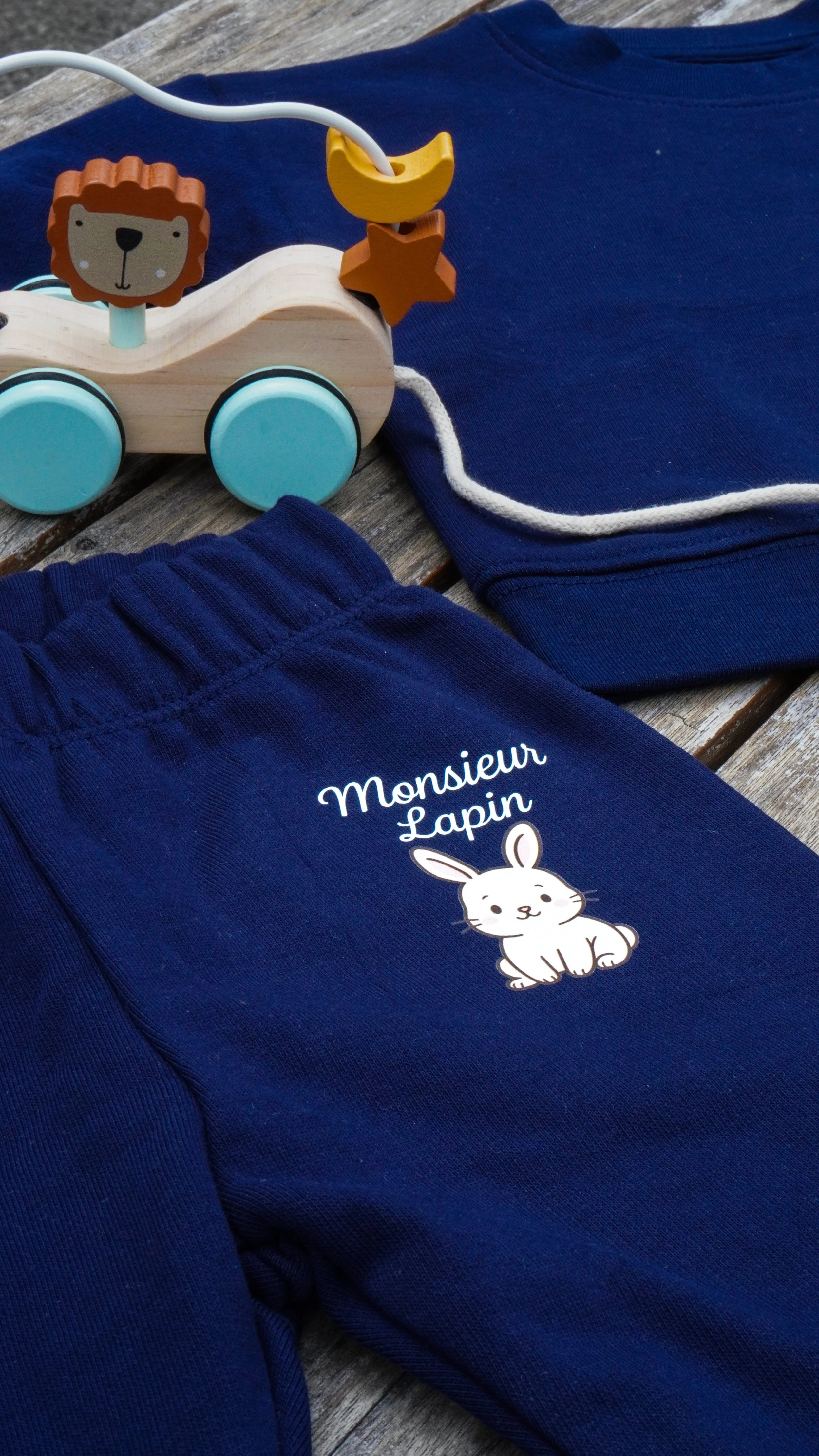 Vêtements bleus pour enfant avec un dessin de lapin et la mention "Monsieur Lapin" ainsi qu'un jouet en bois représentant un lion, une étoile et une lune.