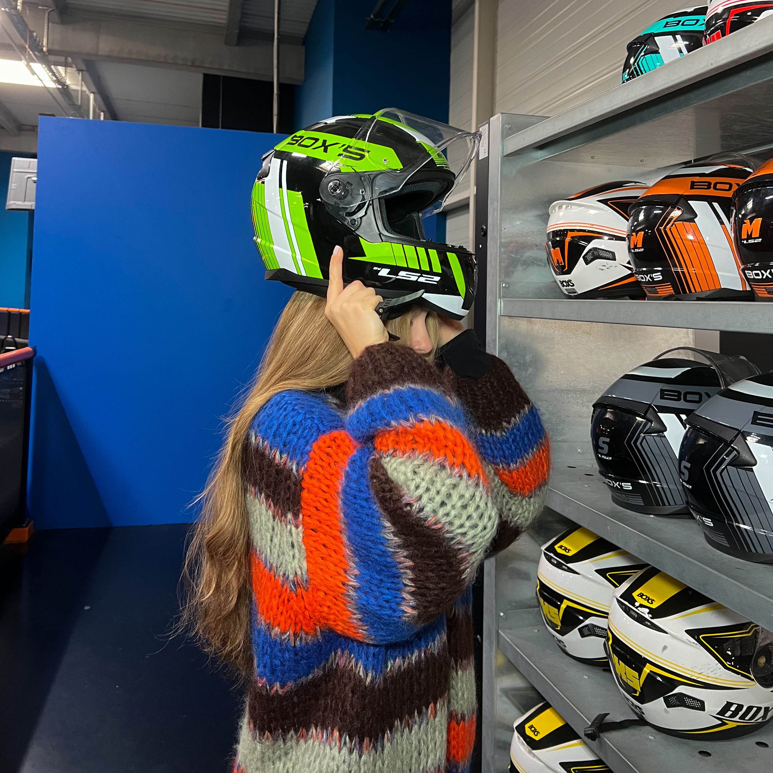 Une femme essaie un casque de moto noir, vert et blanc dans un magasin spécialisé. Plusieurs autres casques sont exposés sur une étagère à côté.