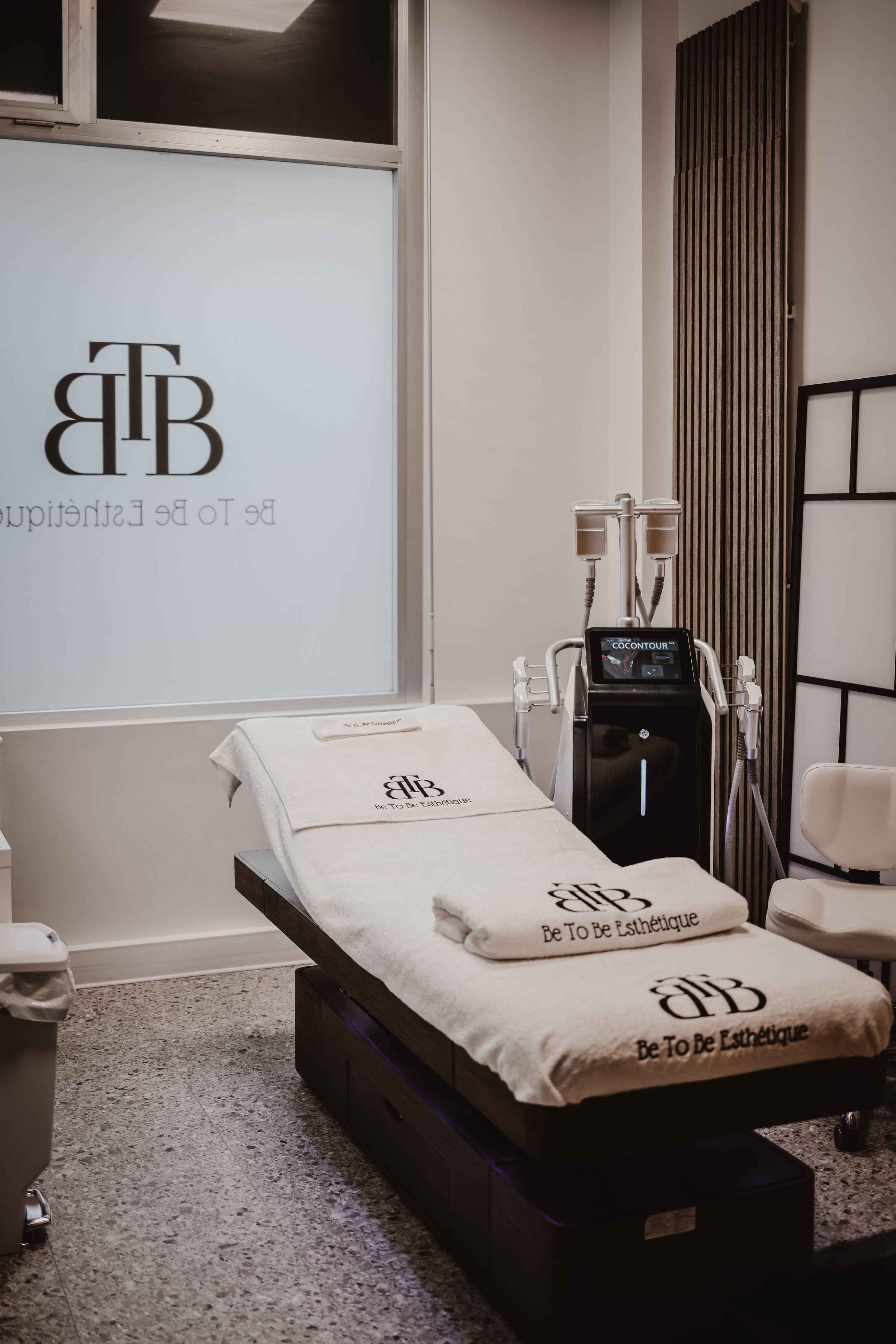 Chambre de soins esthétiques avec table, serviettes, machine COCONTOUR, logo 'Be To Be Esthétique' sur la table et la vitre.
