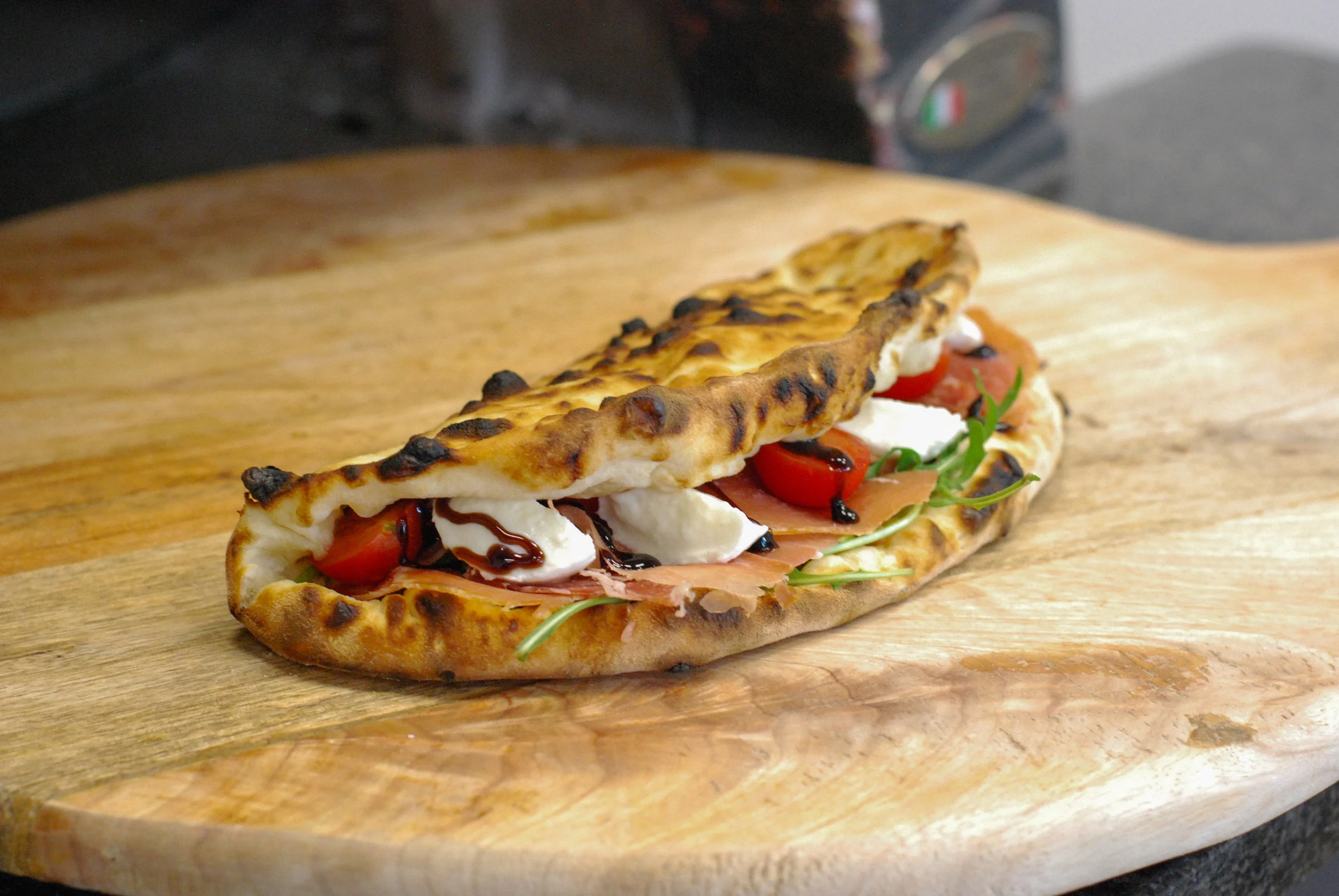 Sandwich avec pain naan, tomates cerises, mozzarella, jambon, roquette, et sauce balsamique sur une planche en bois.