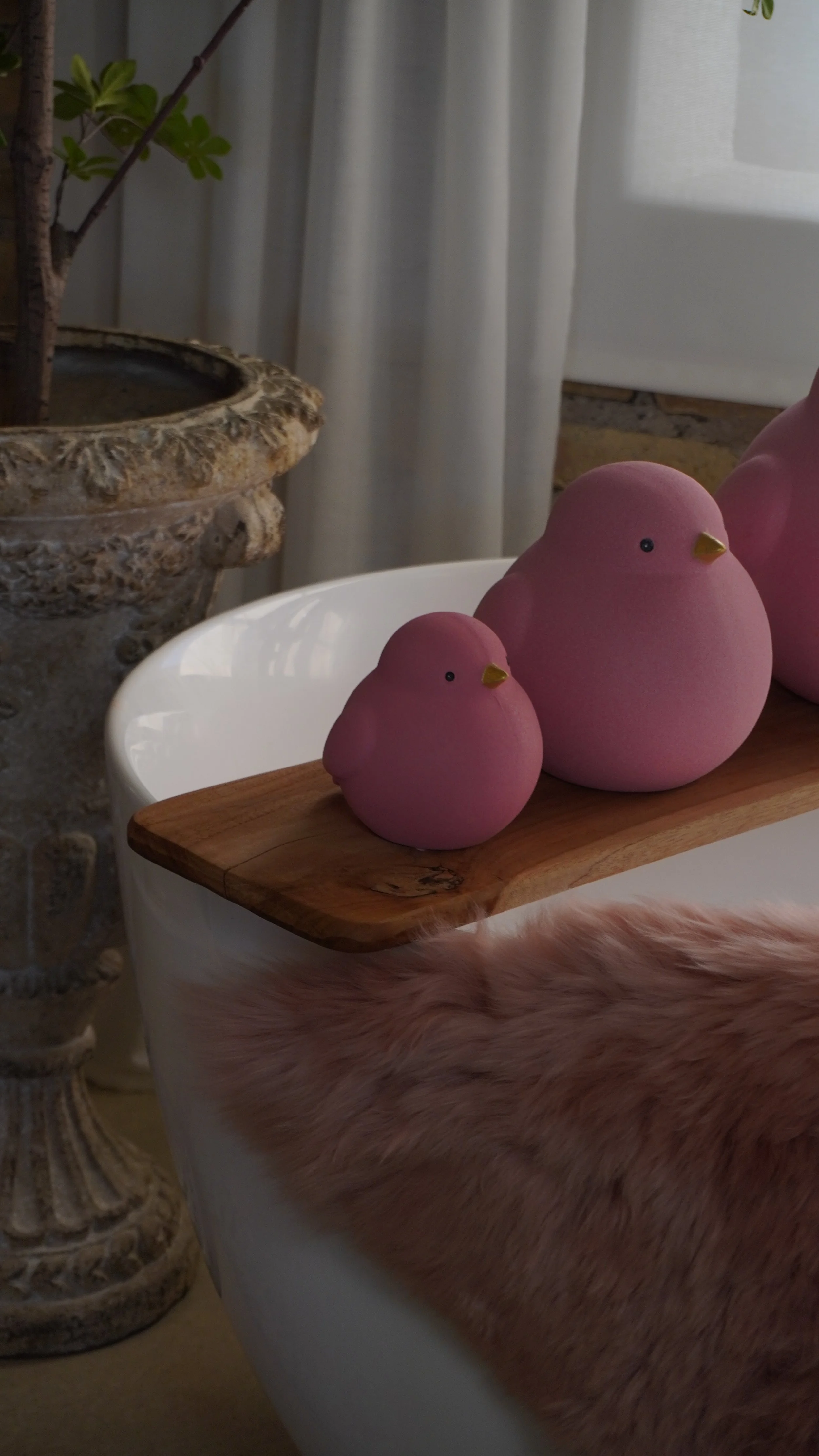 Deux décorations en forme d'oiseaux roses sur une planche en bois posée sur une baignoire blanche, avec une chaise en fourrure rose pâle devant.