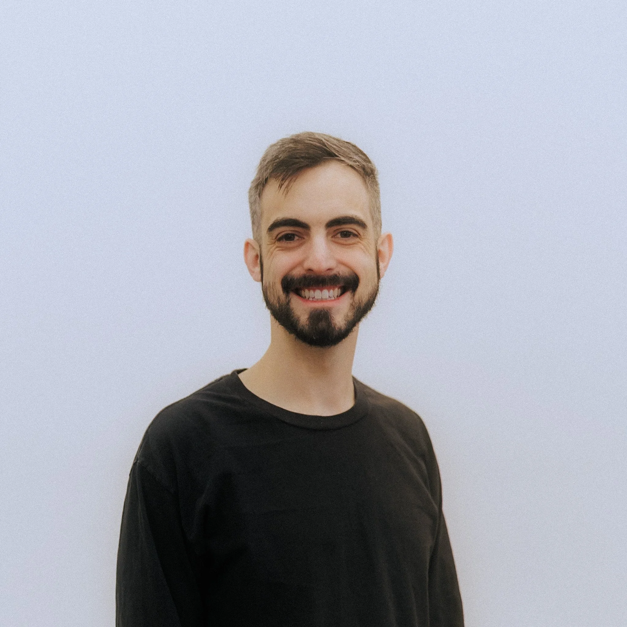 Close-up of a smiling young man with short brown hair, beard, wearing a black shirt, against a plain light background.