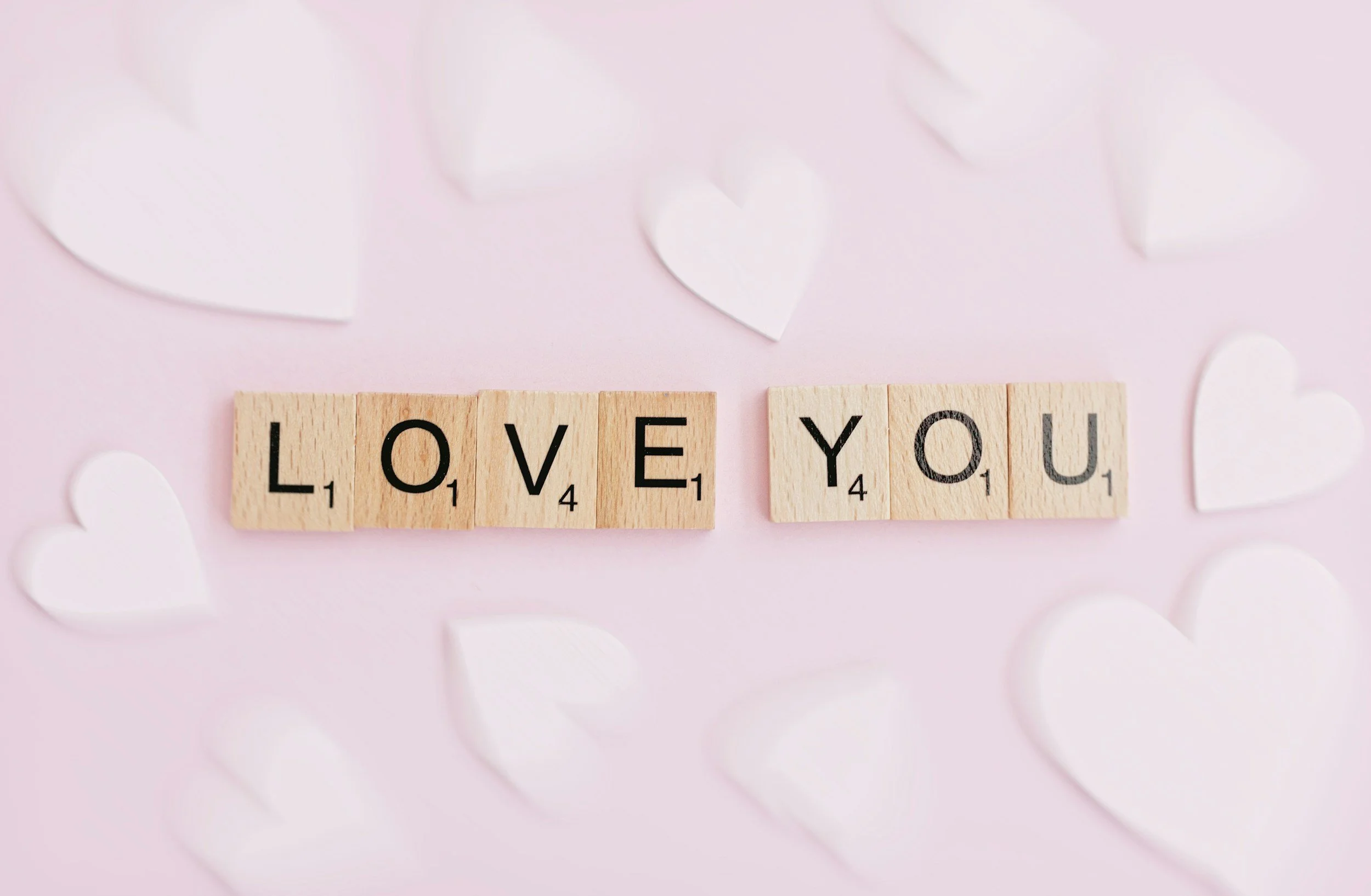 Scrabble tiles spelling 'LOVE YOU' on a pink background with white paper hearts scattered around.