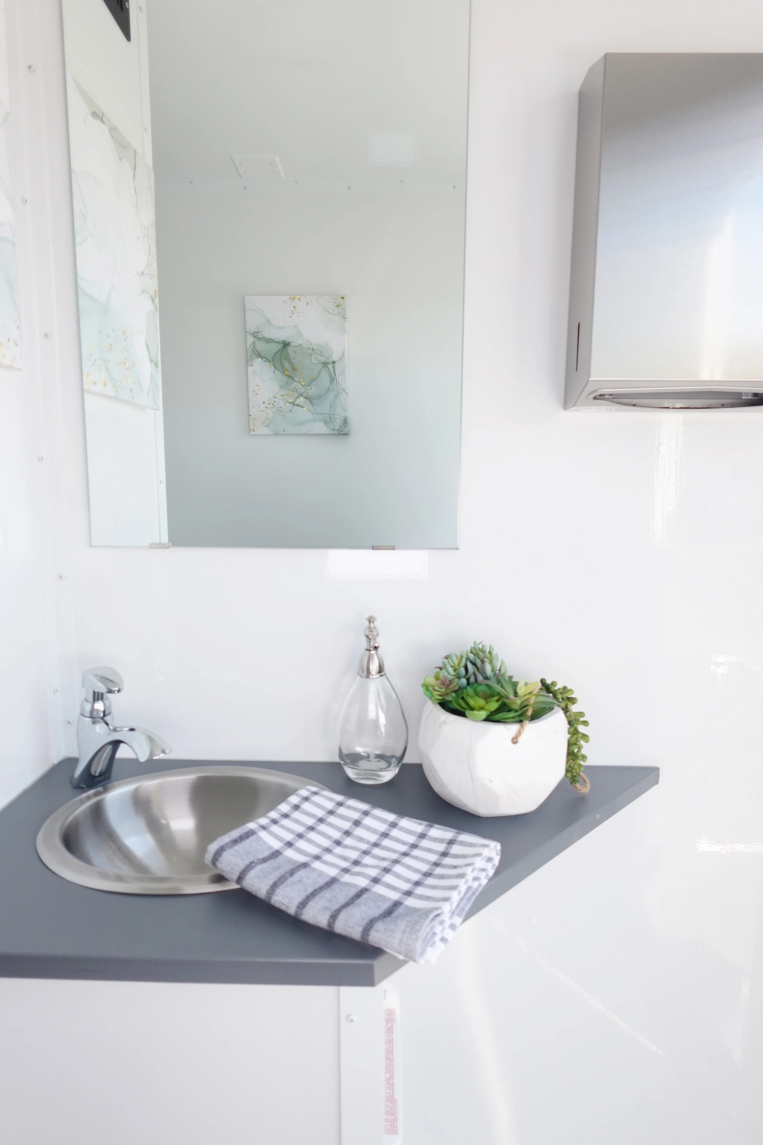 A small bathroom sink area with a round metallic sink, a gray countertop, a checkered towel, a glass soap dispenser, a white planter with succulents, and a mirror reflecting abstract artwork.