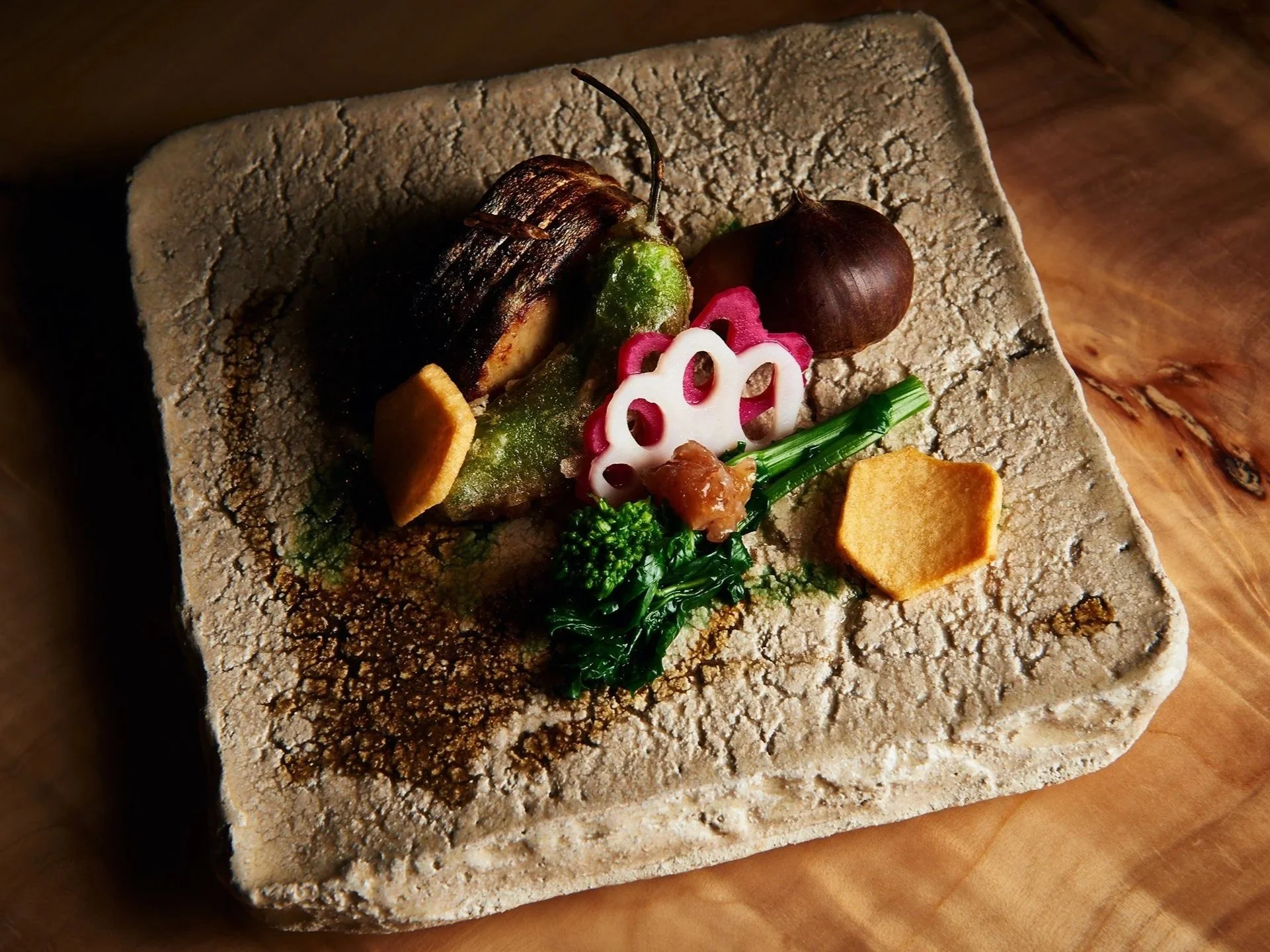 A stone slab with a variety of garnishes, including a cherry tomato, green peppers, pink and white rice cakes, and green vegetables, garnished with sauce, on a wooden surface.