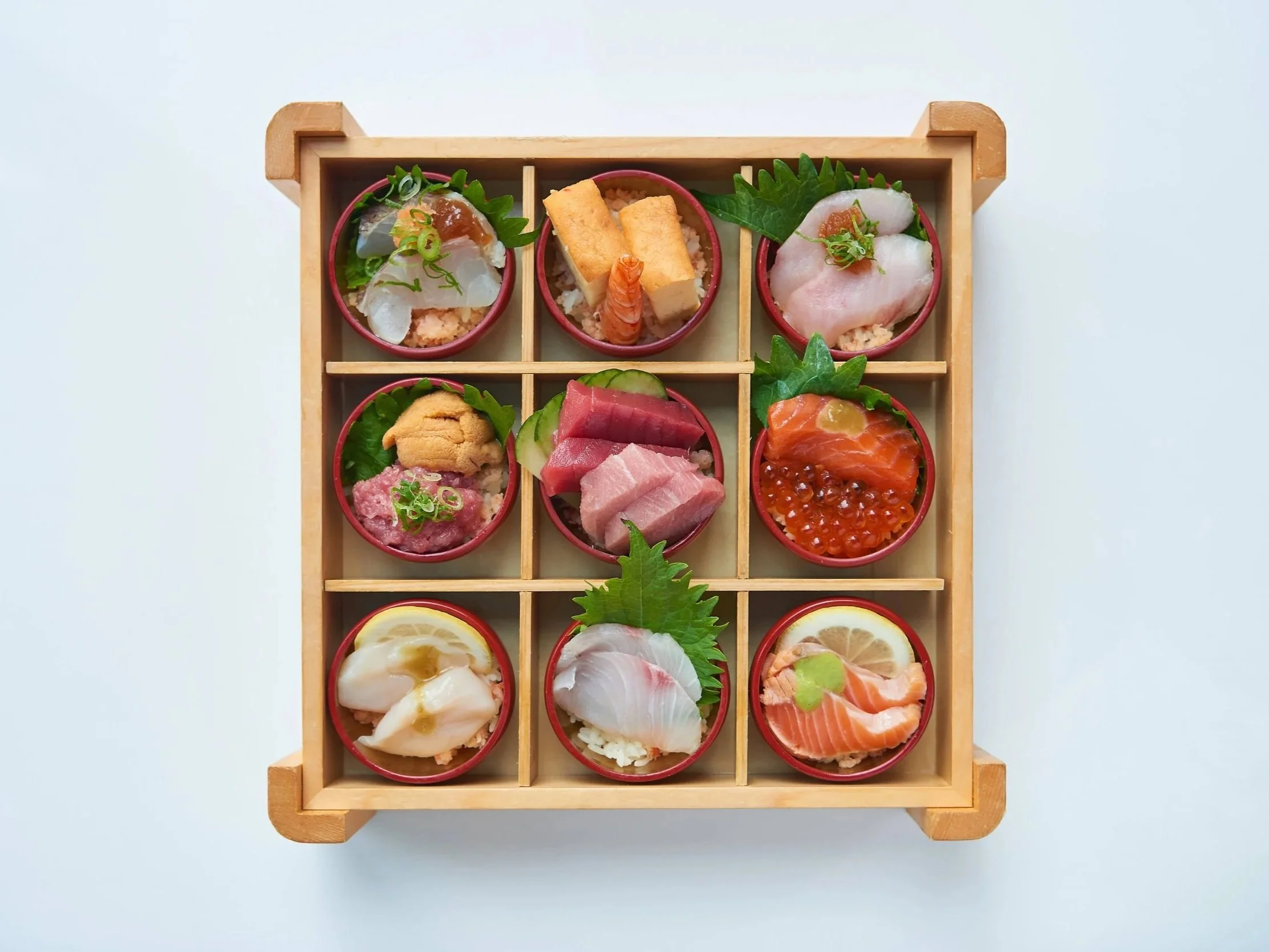 Top view of a wooden bento box with nine compartments containing assorted sashimi, sushi, and garnishes.