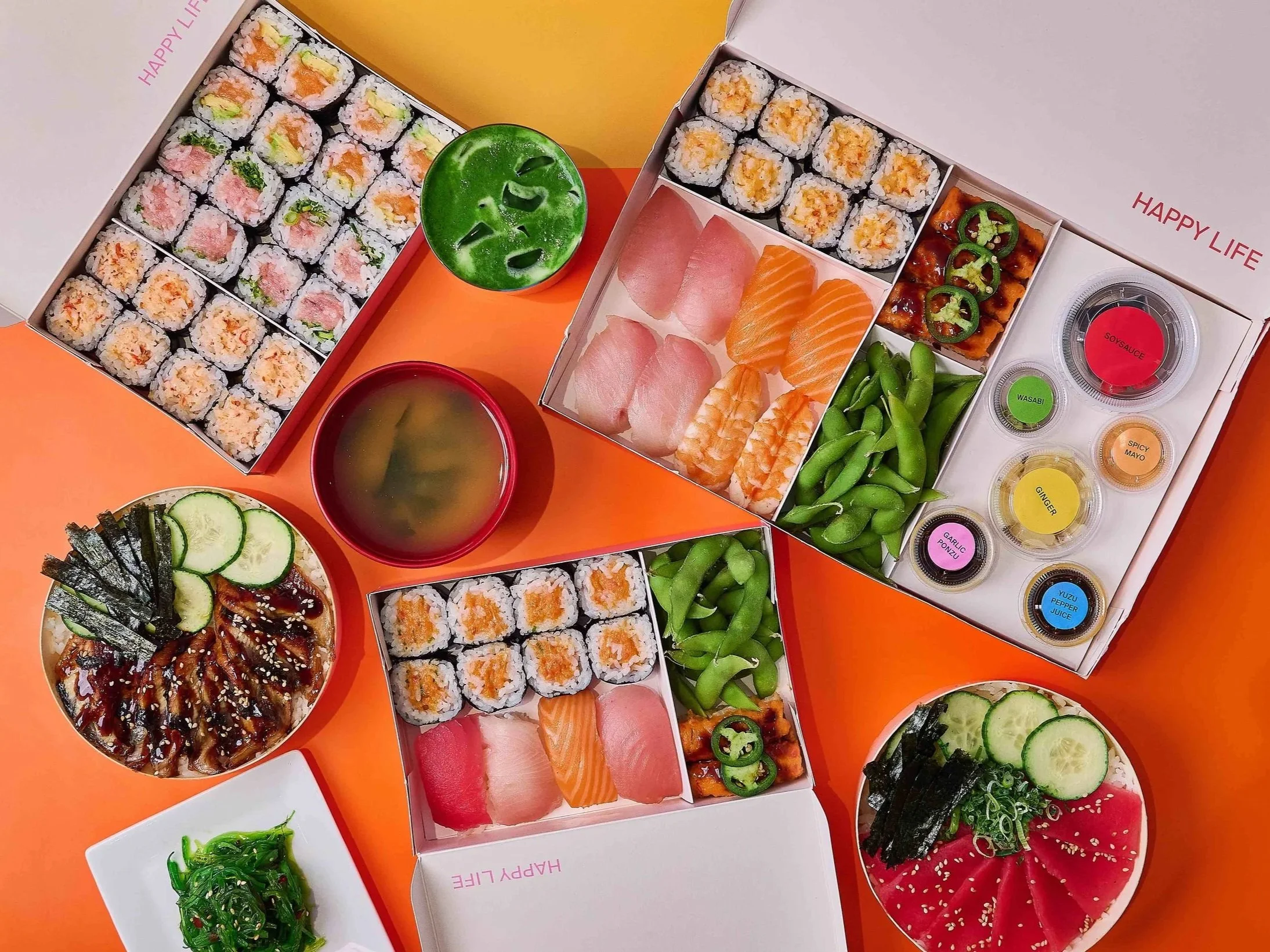 Assorted sushi rolls, sashimi, sashimi with jalapeño slices, edamame, seaweed salad, cucumber slices, and dipping sauces, all placed on an orange surface.