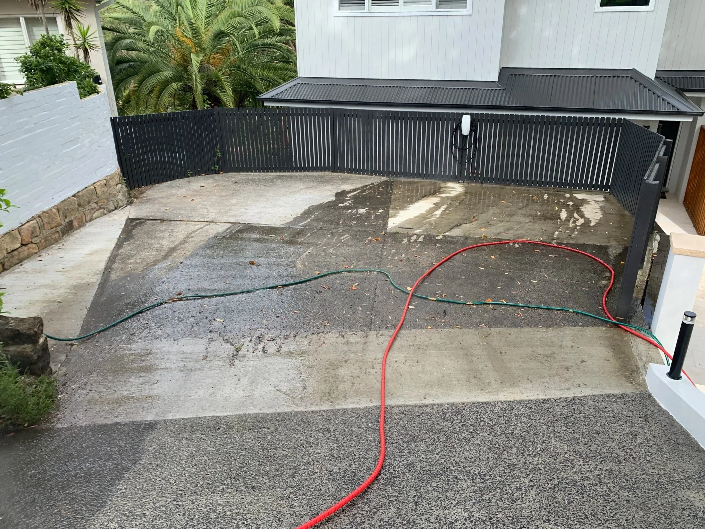 Wet concrete driveway with hoses, partially cleaned, in front of a modern house with black fence and tropical plants.