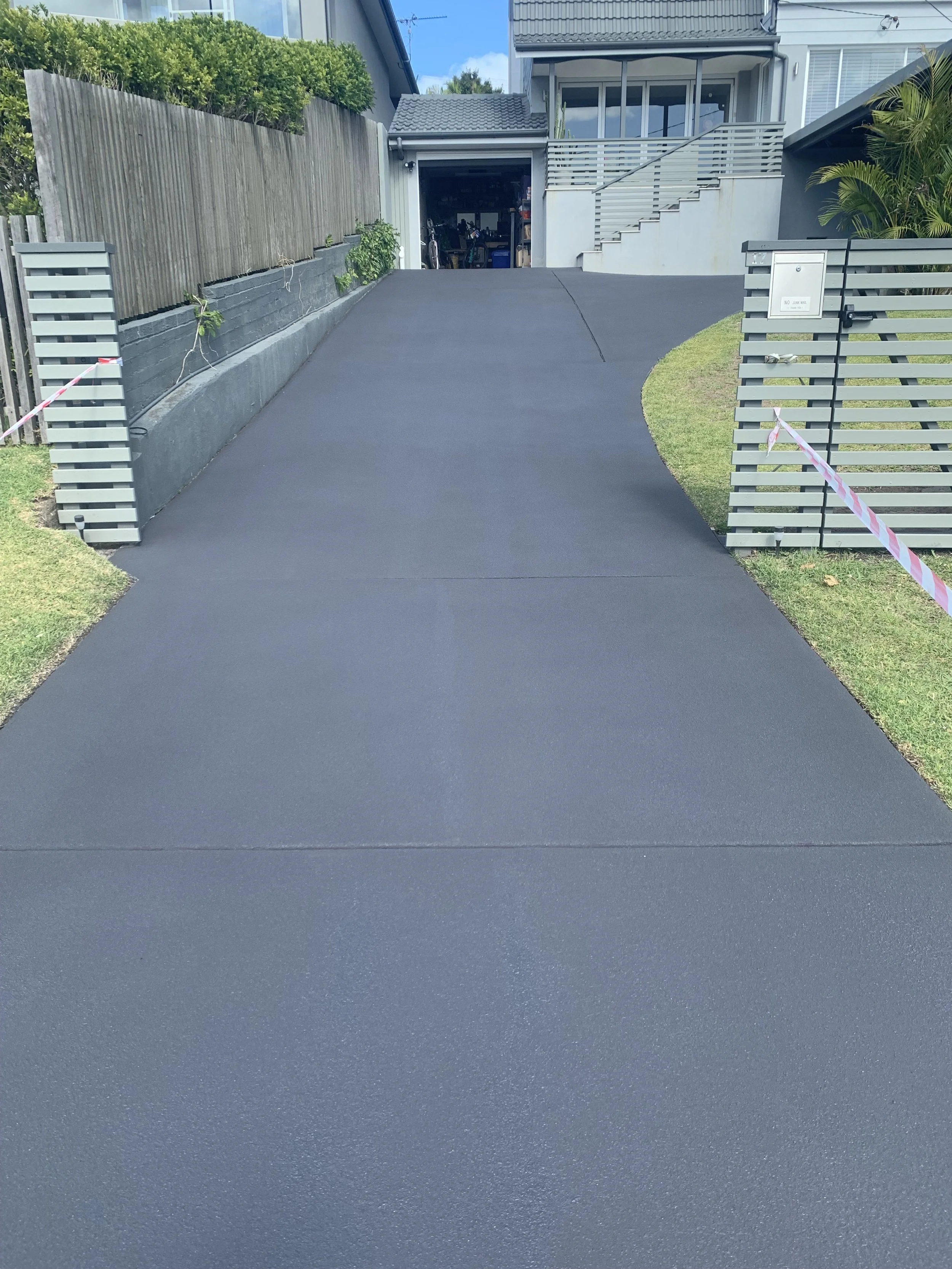Newly paved black asphalt driveway leading to a garage with open doors, flanked by modern gray fencing and green landscaping.