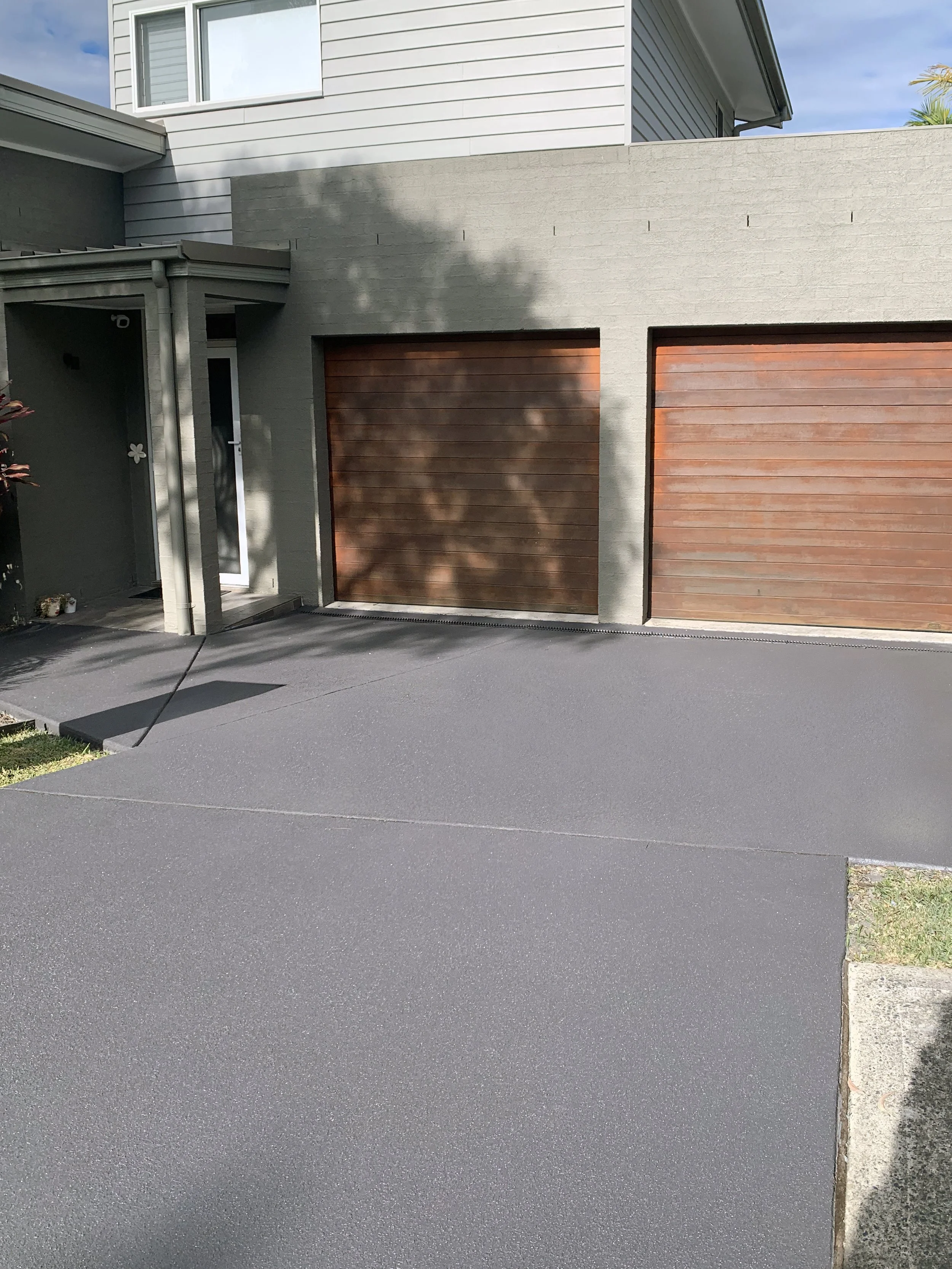 Front view of a residential house with two garage doors, one dark brown and one lighter brown, a side entrance door, and a freshly paved black driveway.