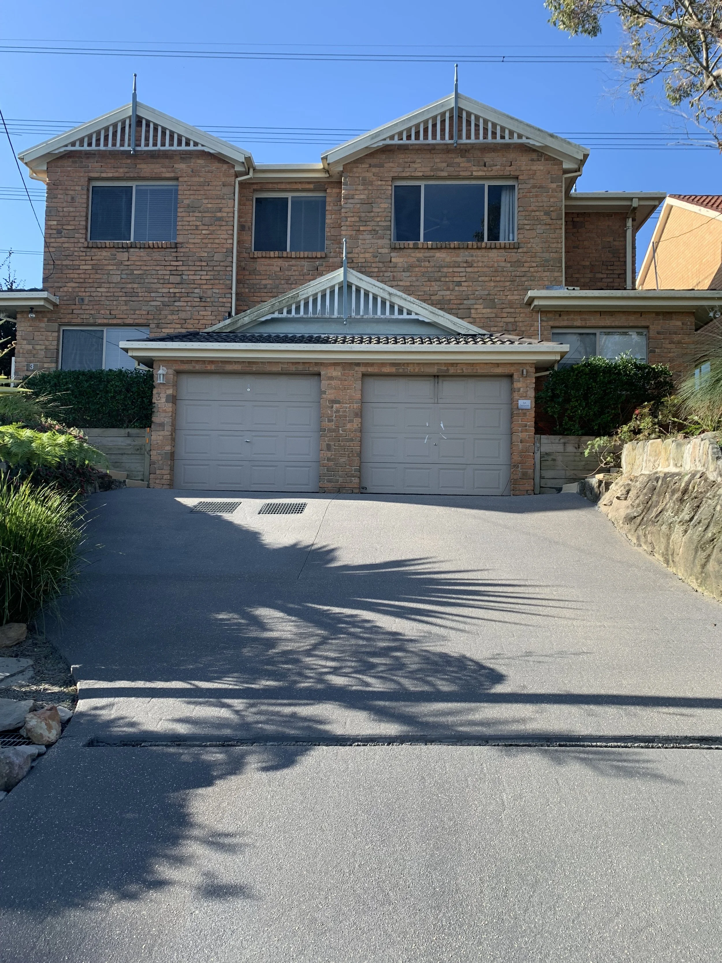 A two-story brick house with a double garage, situated on a sloped driveway. The house has triangular gables and windows on both floors, with a clear blue sky above and a tree casting shadows on the driveway.