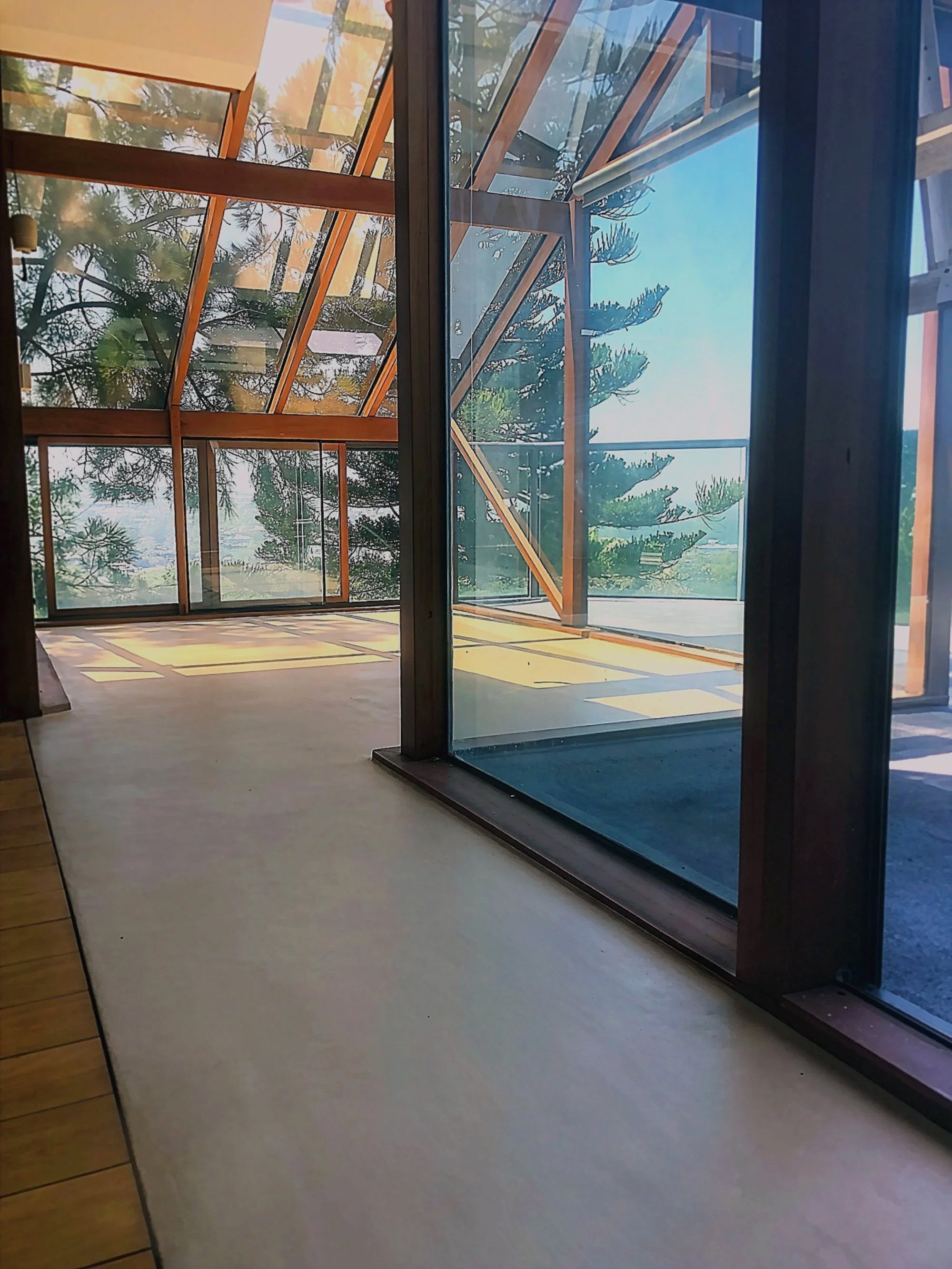 Interior view of a modern house with large glass windows and wooden frames, overlooking trees and a clear sky outside.