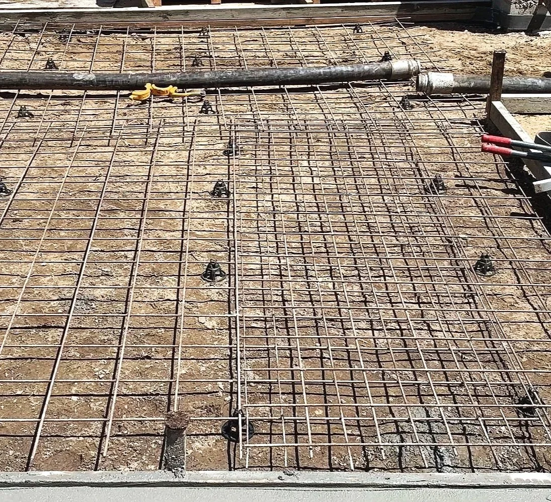 Construction site showing steel rebar framework for concrete slab, with foundation pipe and construction tools.
