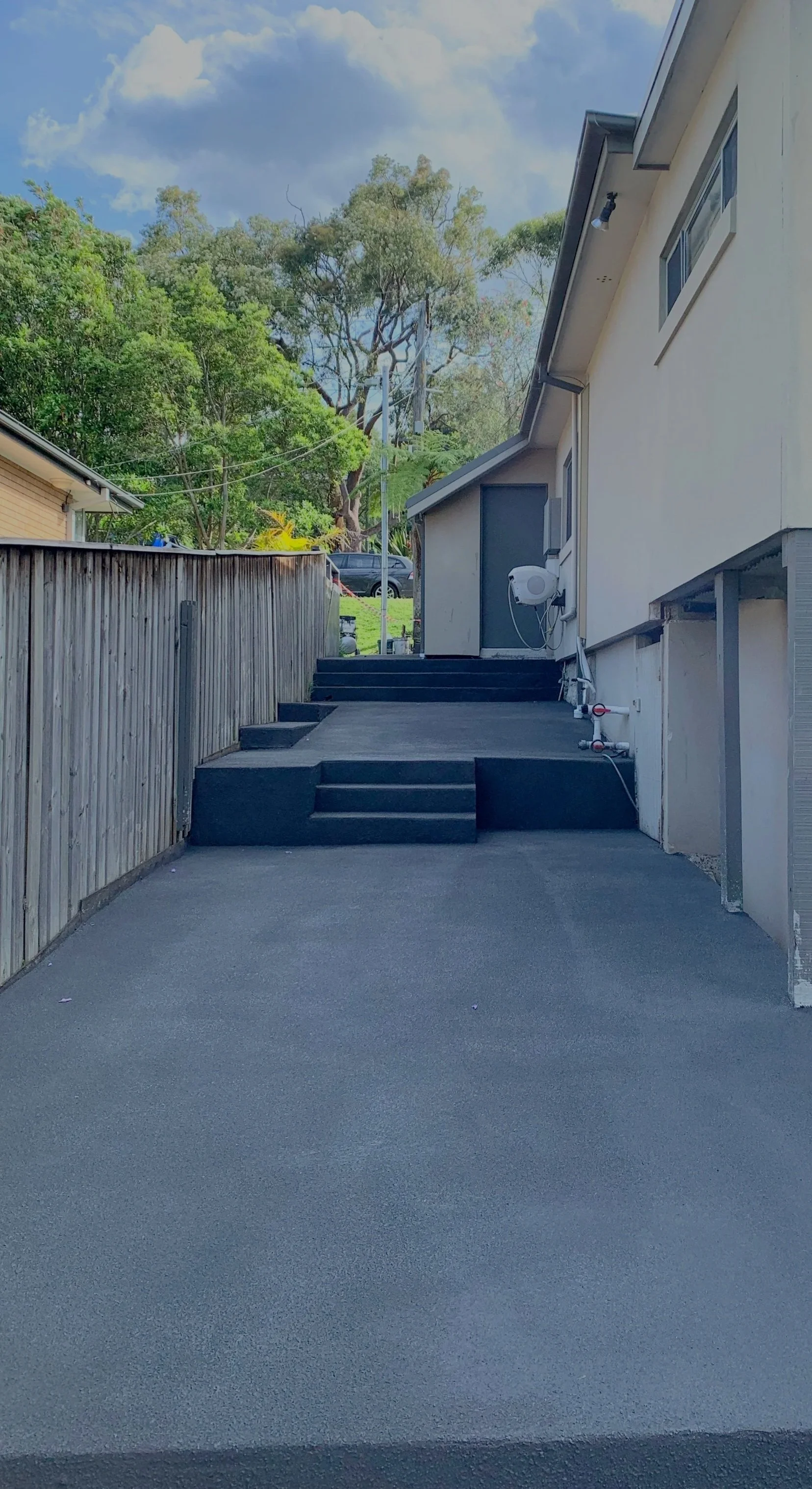 Backyard with newly poured concrete patio, stairs, and an outdoor water heater mounted on the side of the house. Fencing and greenery in the background with trees and a blue sky.