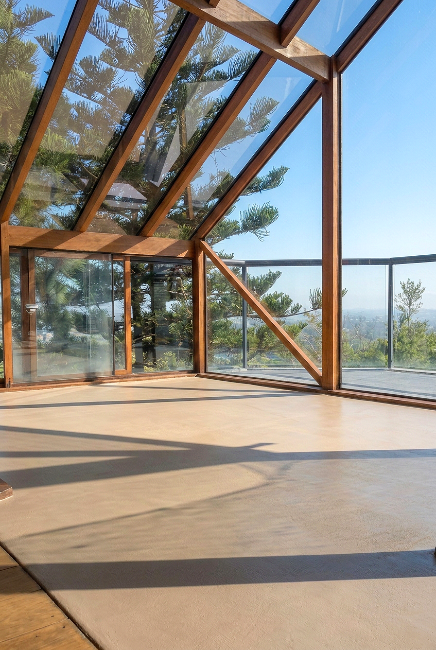 Glenn monckton concrete revival, newly resurfaced sunroom ,Sunlit room with wooden framing and large glass windows showing a view of trees and a clear sky.