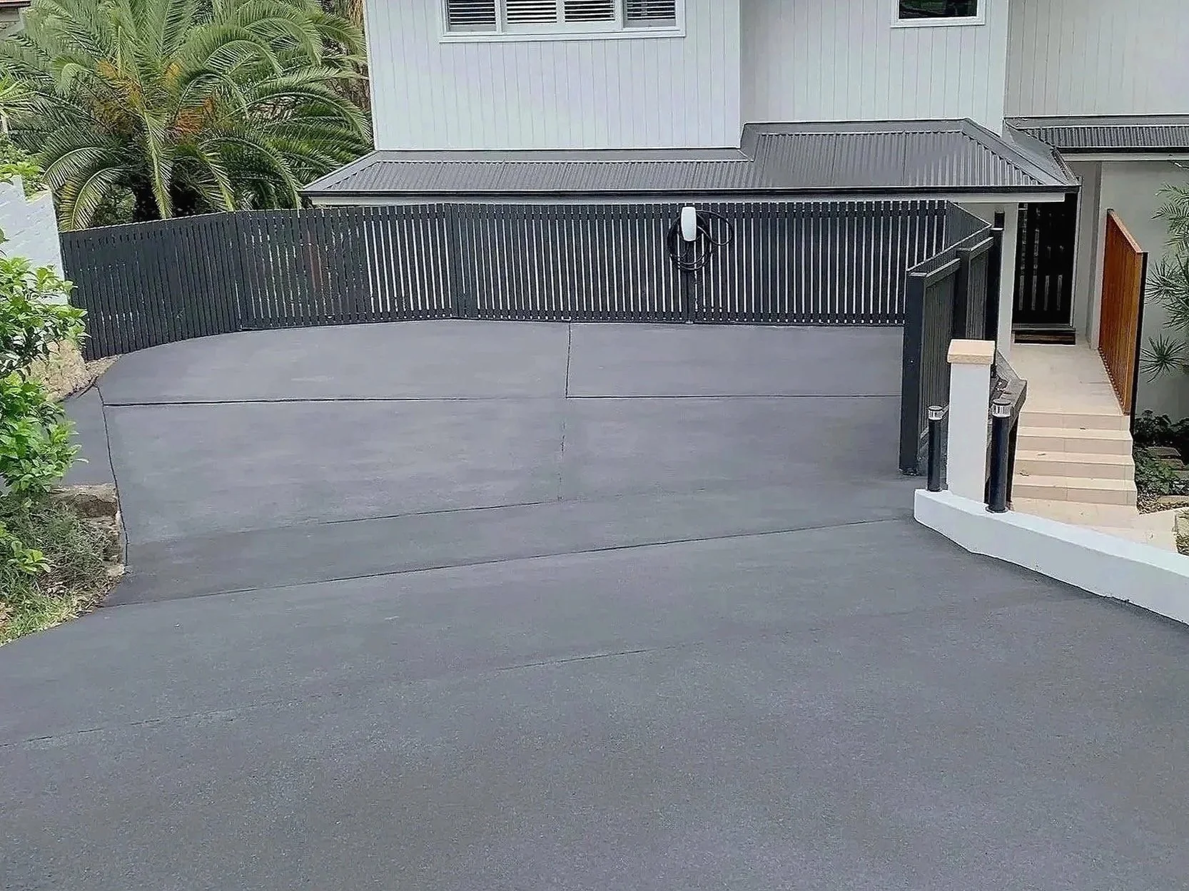Concrete driveway leading up to a modern house, fenced with black vertical slats, with steps on the right side and tropical plants in the background.