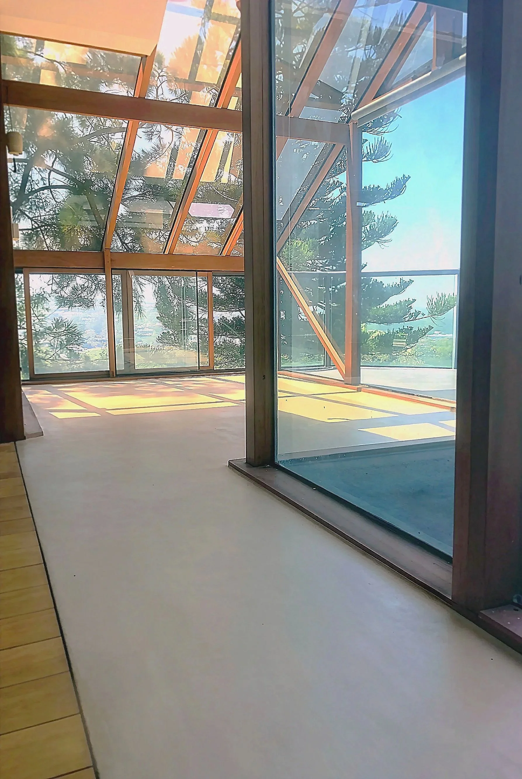 Interior view of a modern house with large glass windows and a wooden framework, overlooking a landscape with trees and a blue sky.