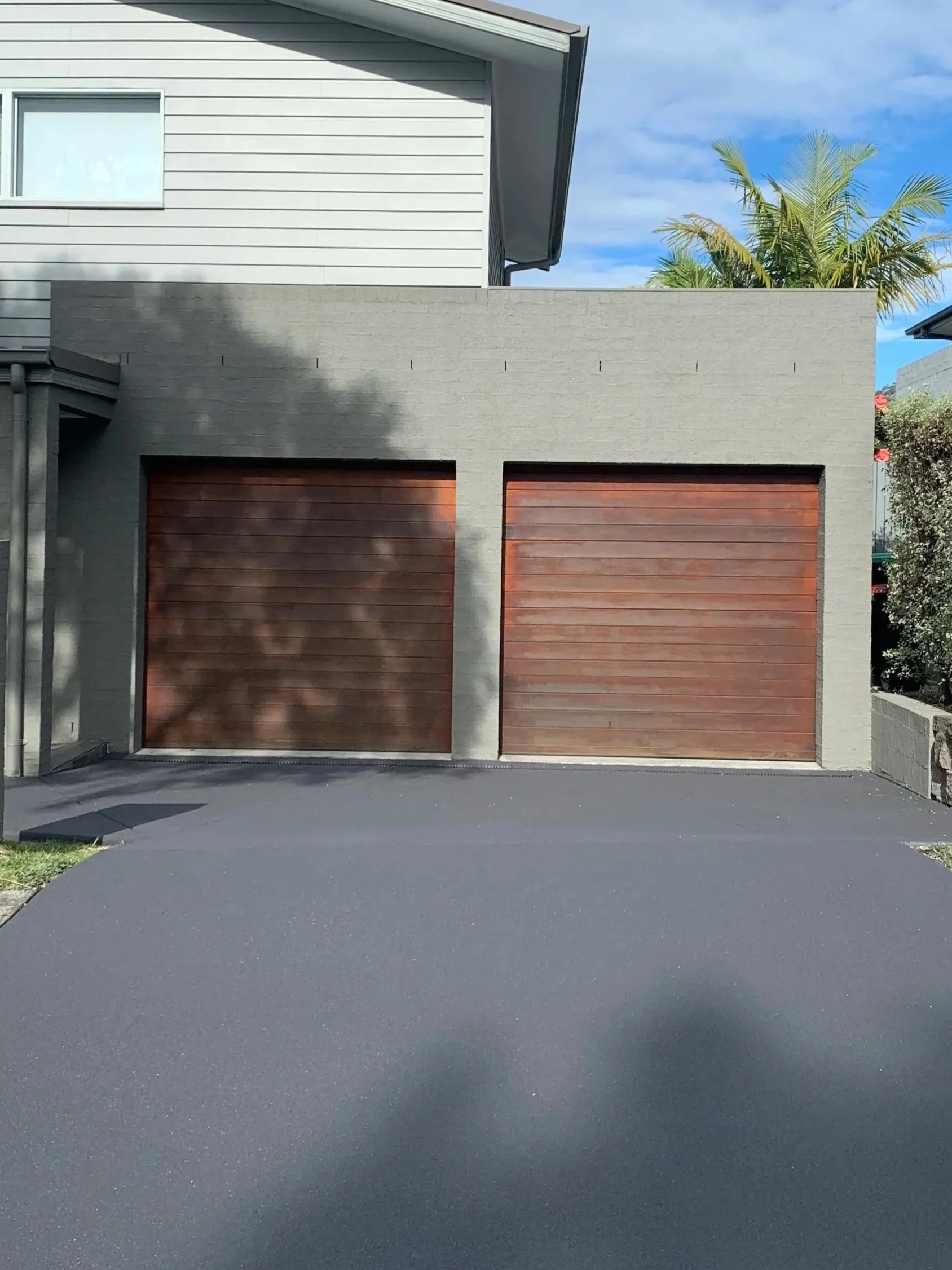 A modern residential building with two wooden garage doors, a gray brick exterior, and a light gray house above with large windows, set against a blue sky with some clouds and palm trees in the background.