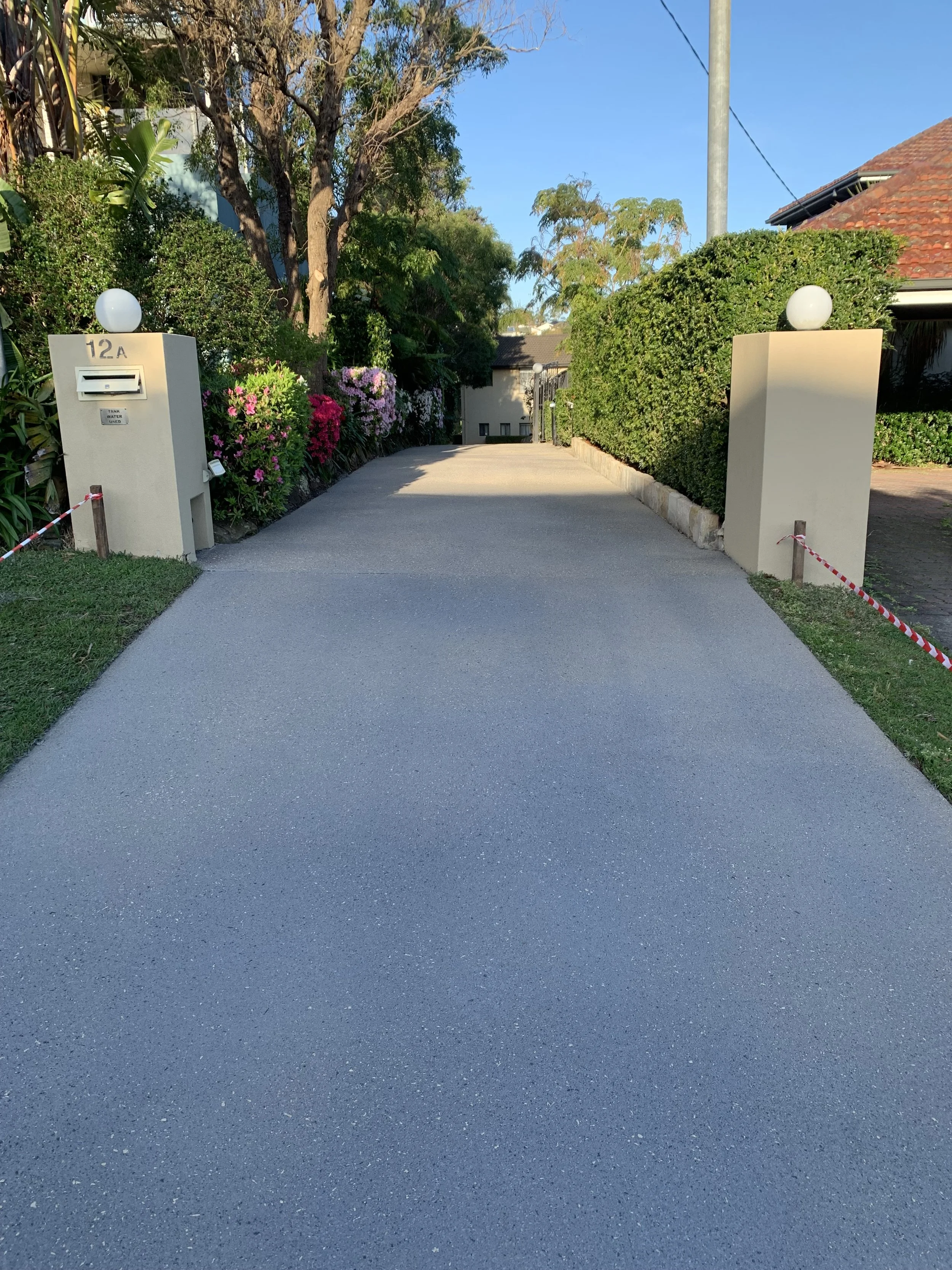A residential driveway with a smooth, light-colored concrete surface, flanked by green grass and bushes on either side. The driveway leads to a house at the end, with trees and clear blue sky overhead.