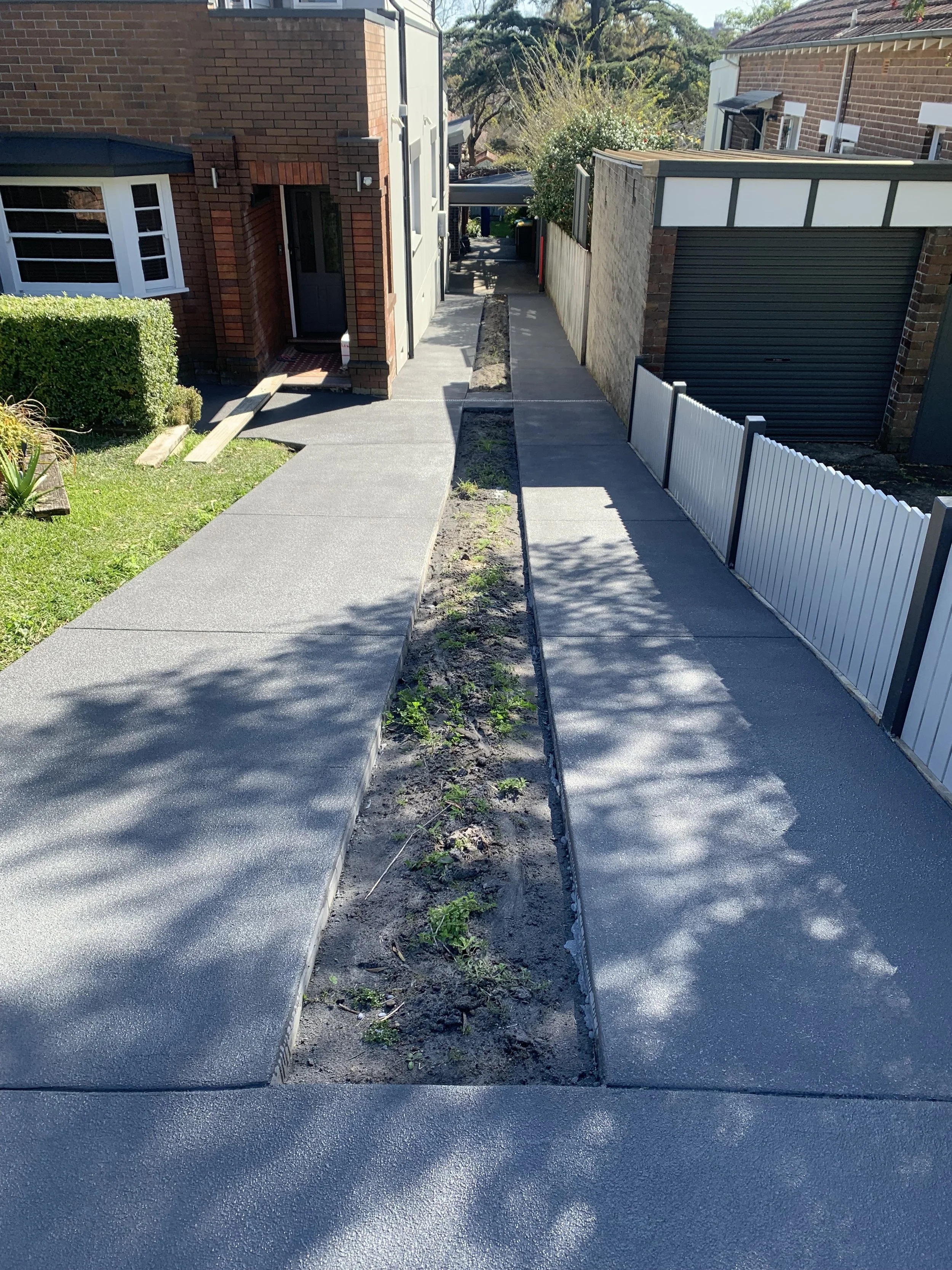 Sidewalk with a narrow garden bed in the middle, adjacent to a house with brick and white exterior. Shadow of tree leaves on the sidewalk, and a white fence on the right.