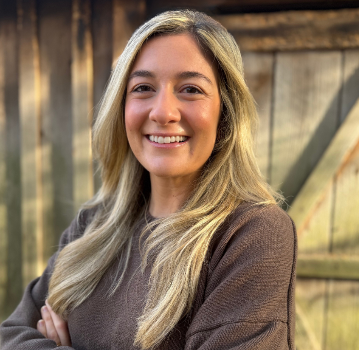 A young woman with long blonde hair smiling outdoors with a wooden fence in the background.