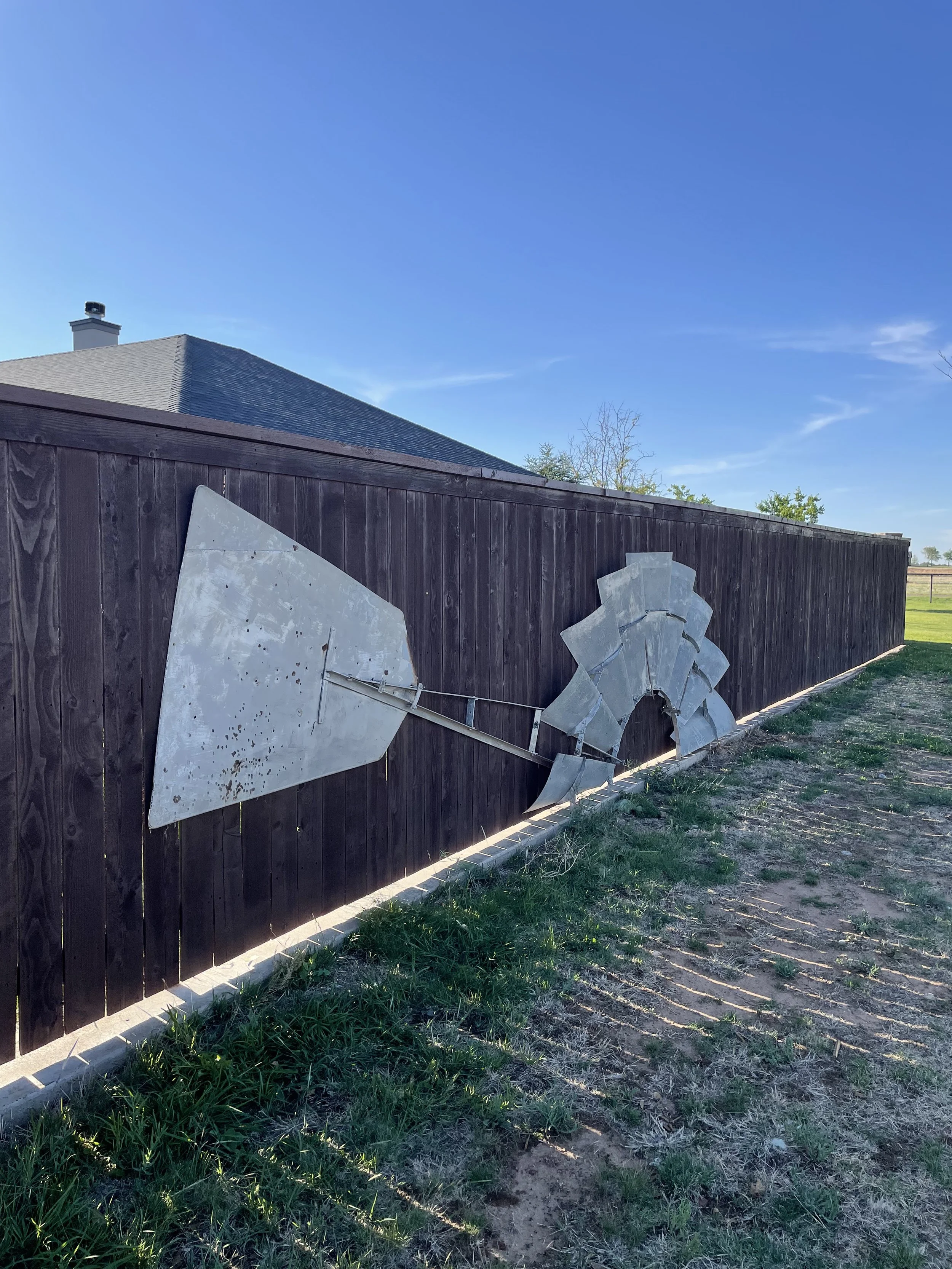 Metal abstract art installation on a dark wooden fence, consisting of geometrical shapes and curved pieces, with a house and trees in the background under a clear blue sky.
