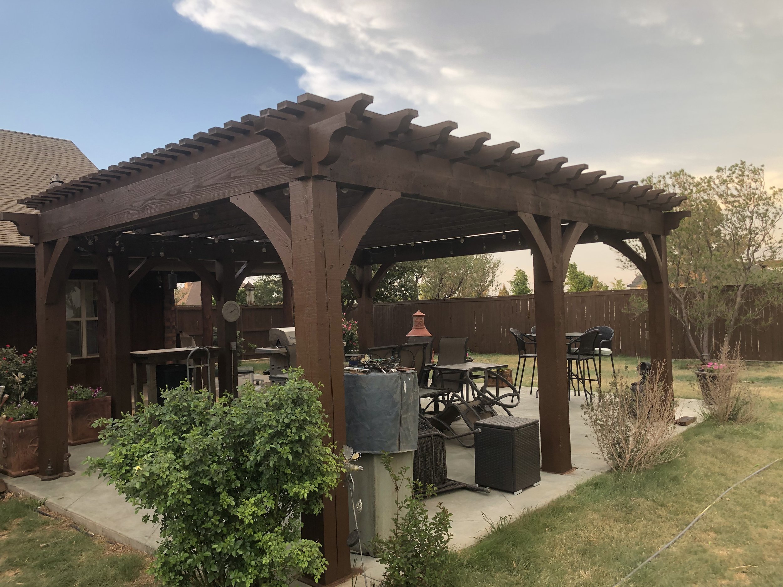 Backyard patio with a wooden pergola, outdoor furniture, and plants, under a partly cloudy sky.