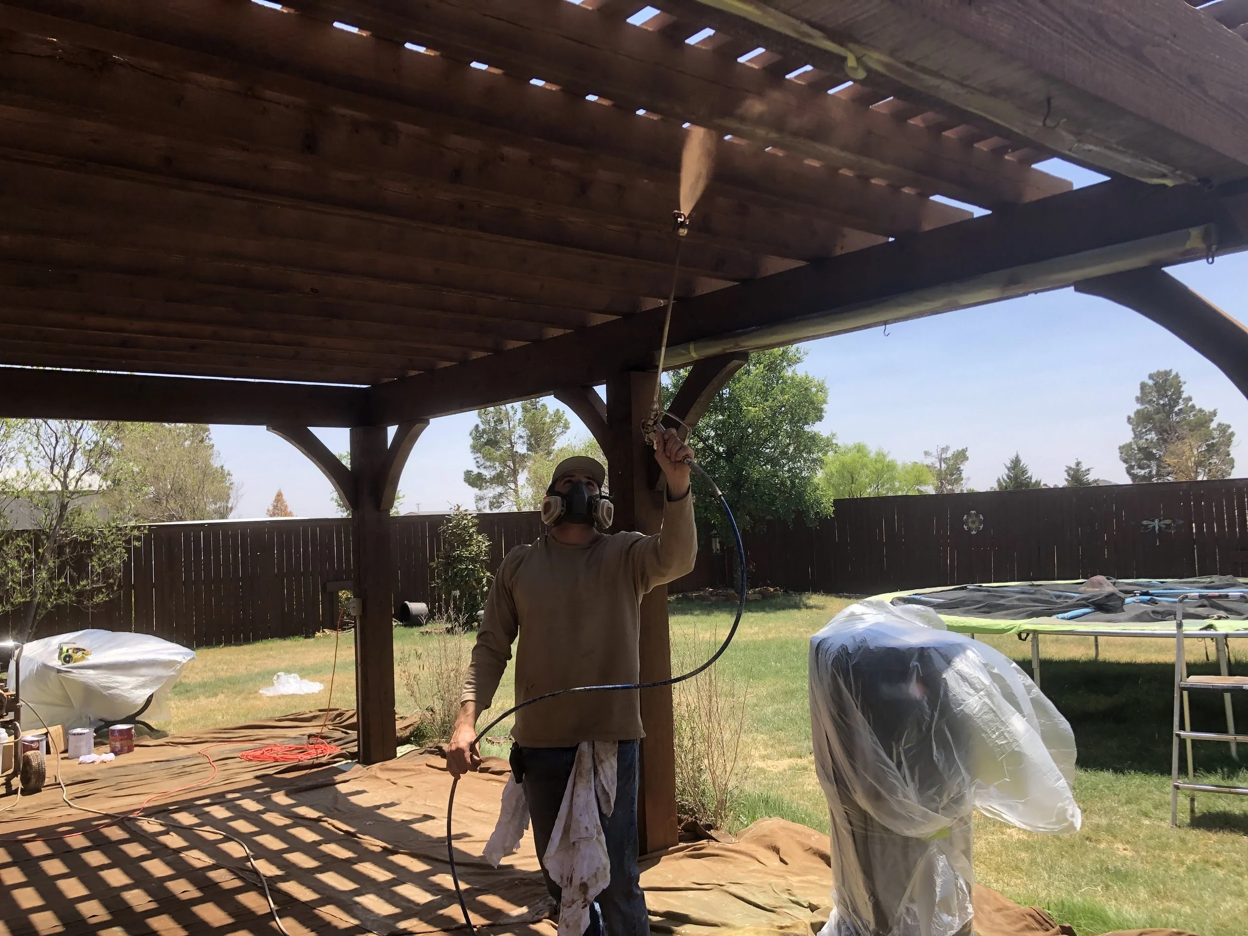 A person wearing a mask and safety gear using a spray gun to paint or stain the underside of a wooden deck in a backyard.