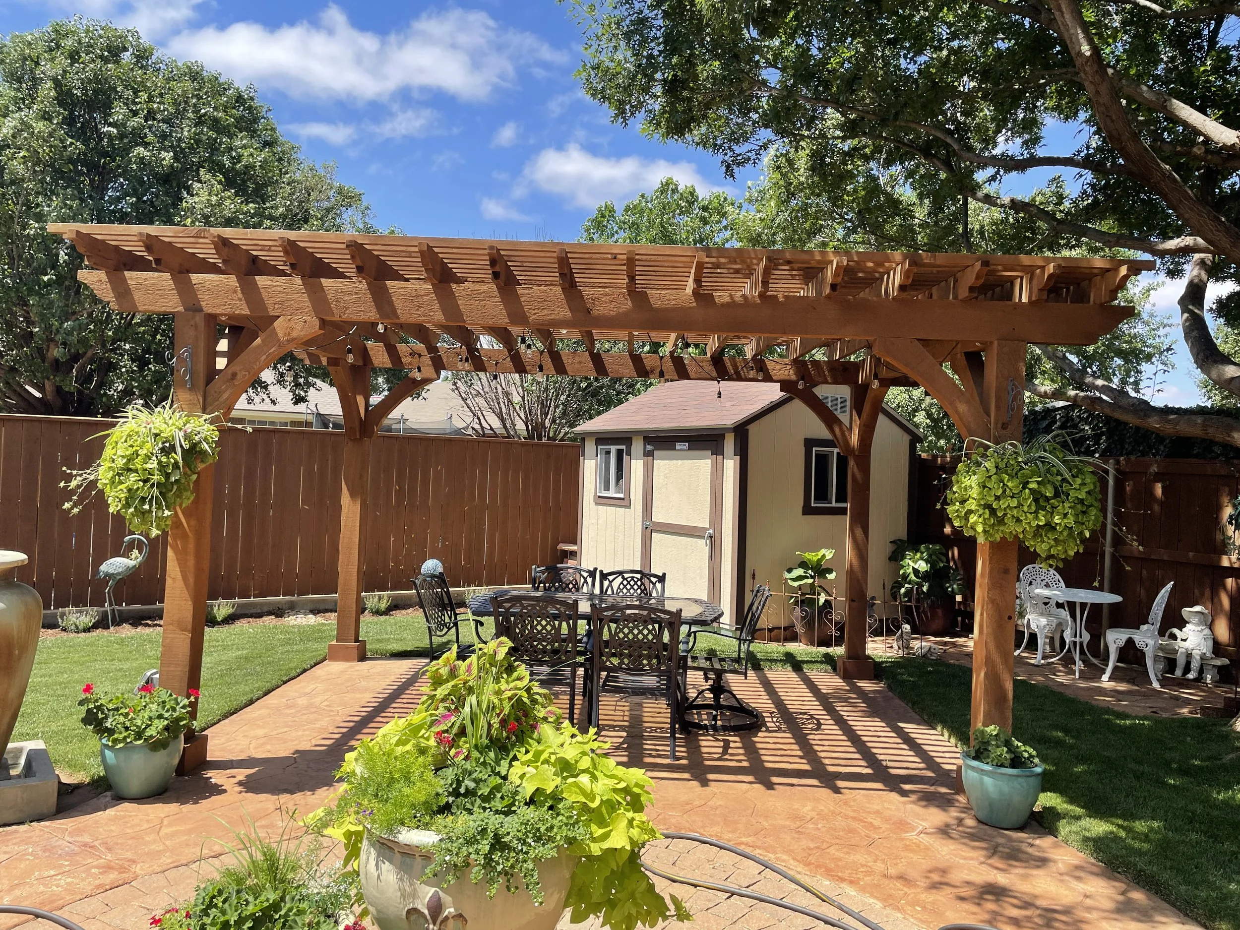 Backyard patio with wooden pergola, black metal table and chairs, potted plants, and a small shed, surrounded by a wooden fence and trees on a sunny day.