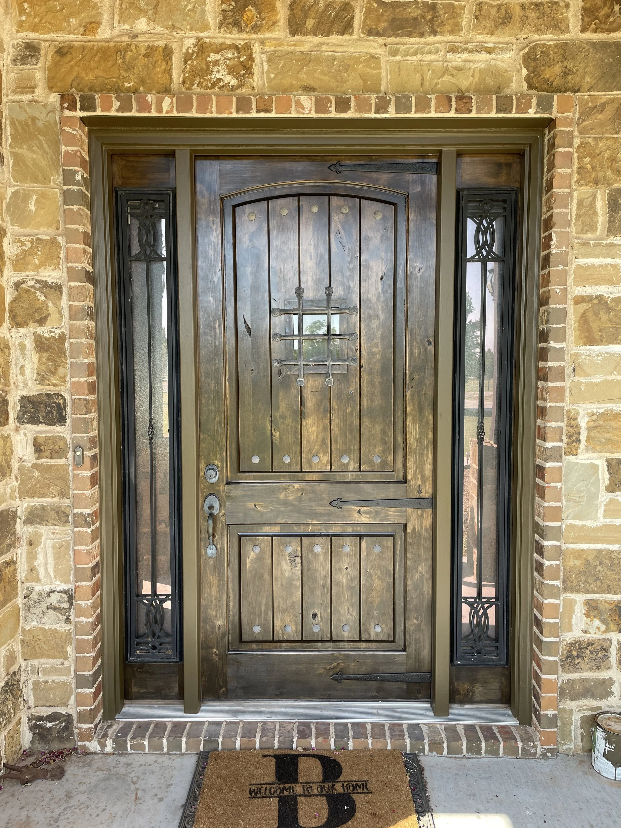 Wooden front door with decorative iron accents, flanked by glass sidelights, set in a brick wall with a welcome mat outside.