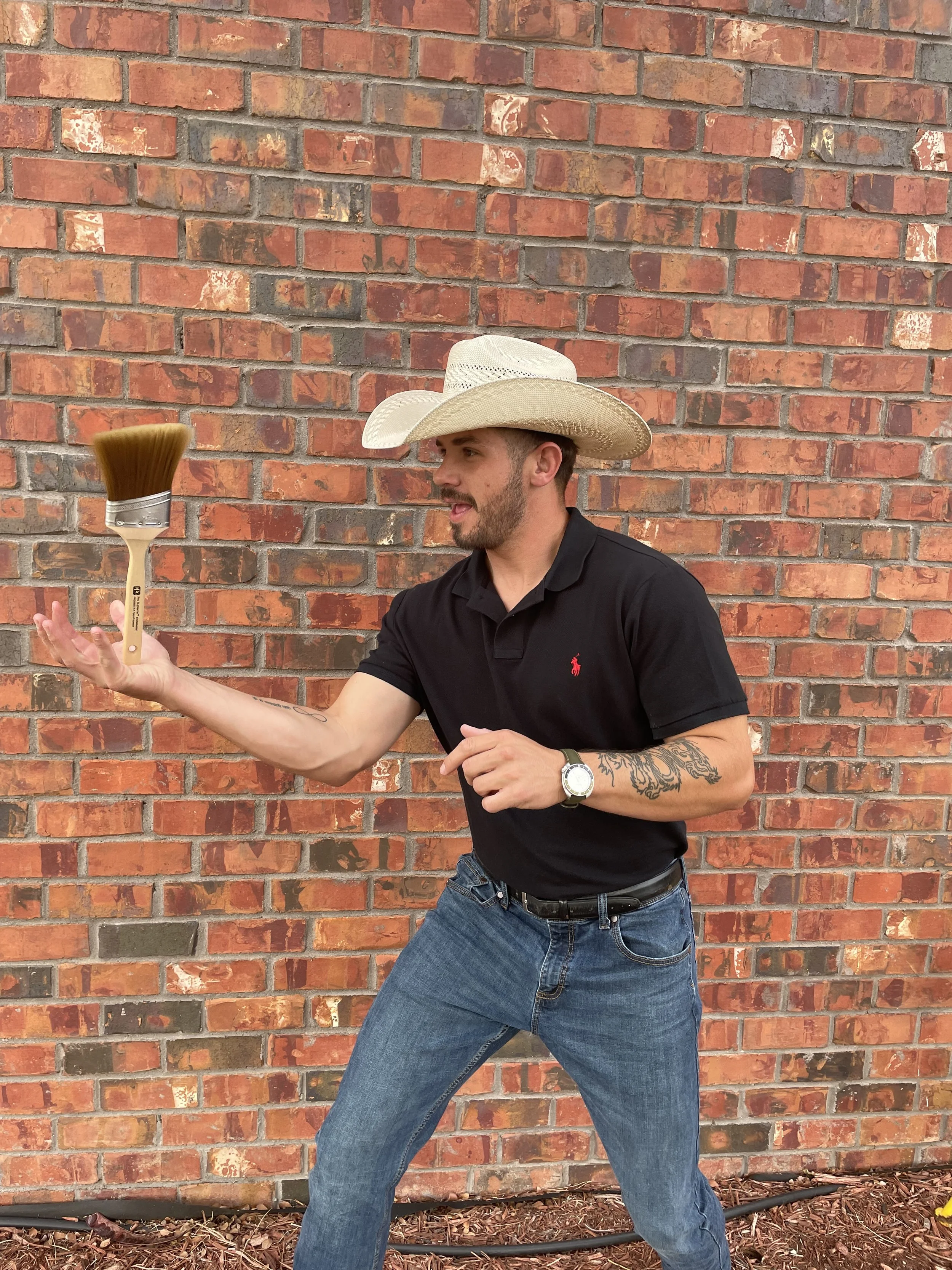 A man wearing a cowboy hat and a black polo shirt with a red logo, holding a paintbrush in his right hand, standing in front of a brick wall.