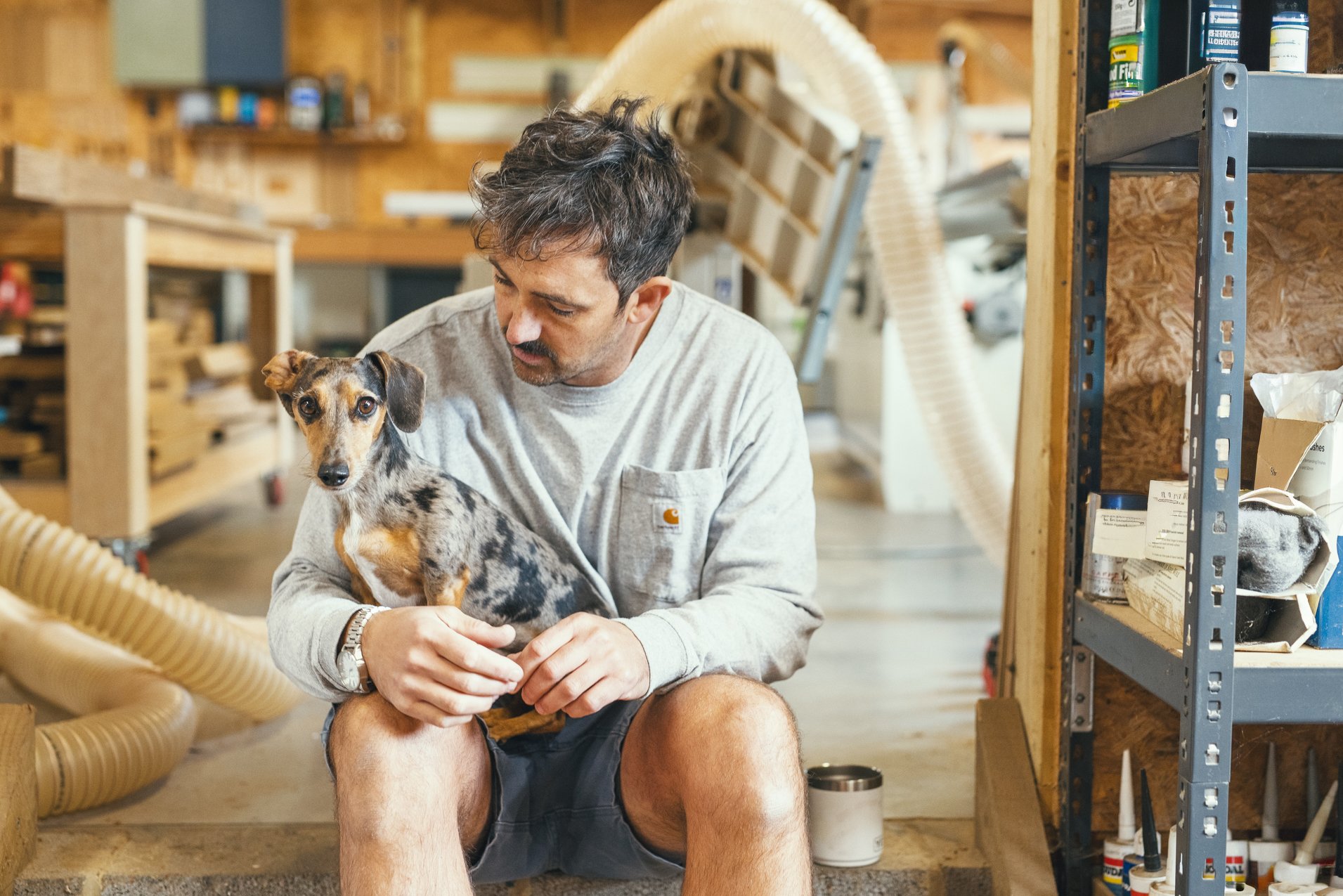 A man is sitting in a workshop holding a small dappled dachshund puppy in his lap, looking at the dog affectionately.