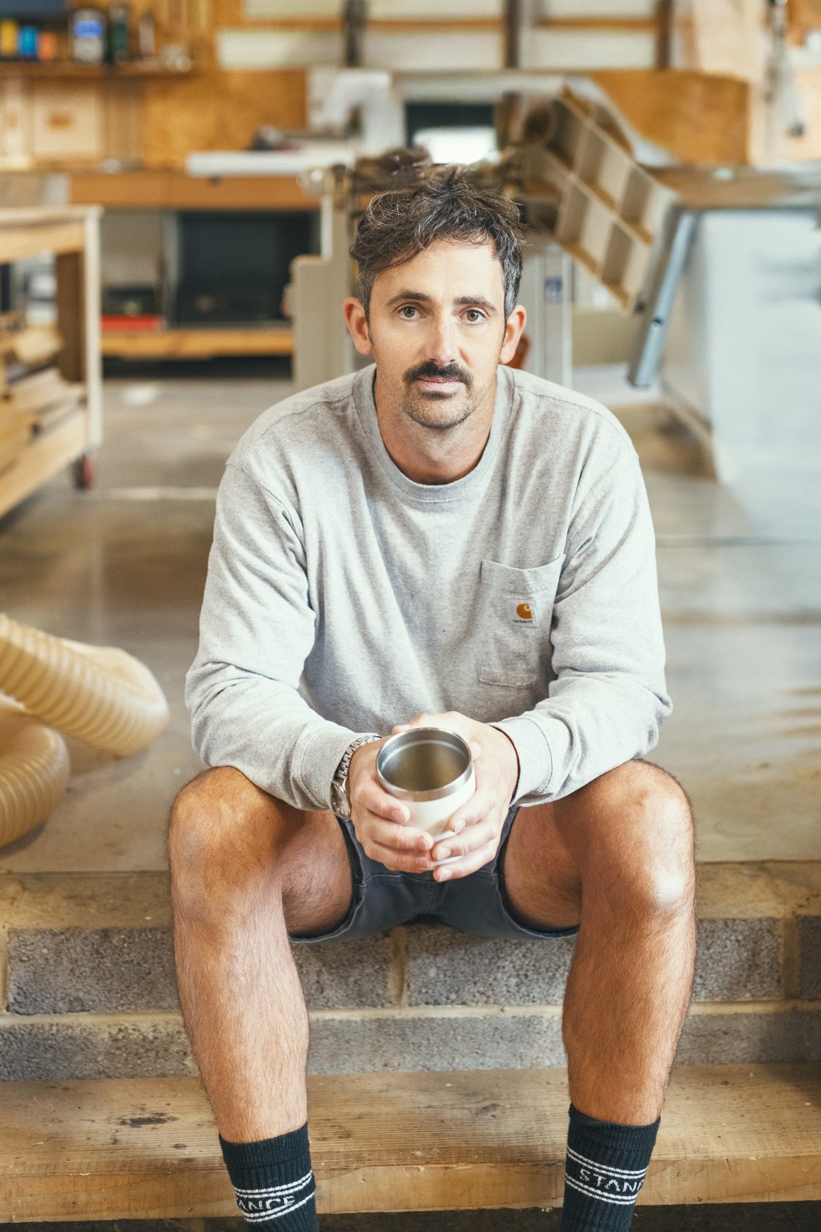 A man sitting on the wooden steps of a woodworking shop, holding a metallic tumbler, with woodworking tools and equipment in the background.