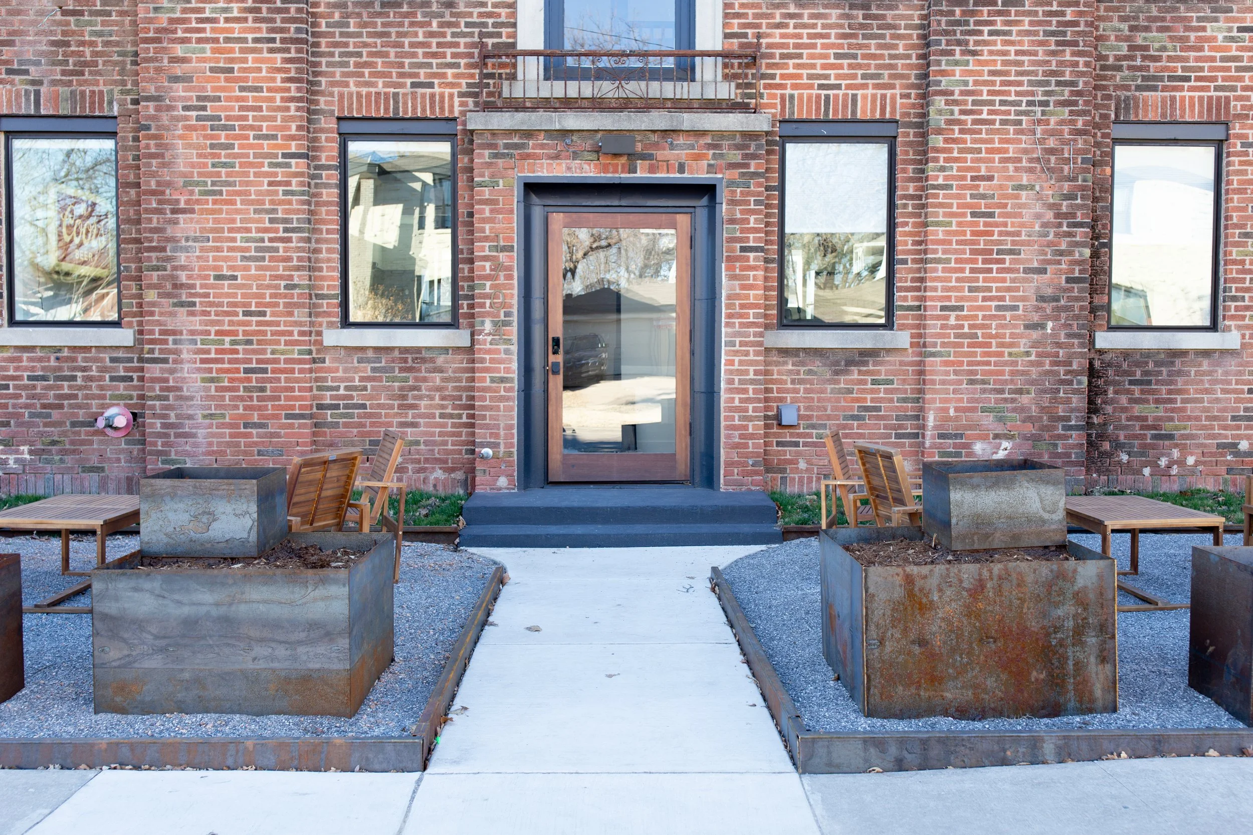Front view of a brick apartment building with a glass door and four large metal planters with soil, outdoor chairs, and a concrete sidewalk.