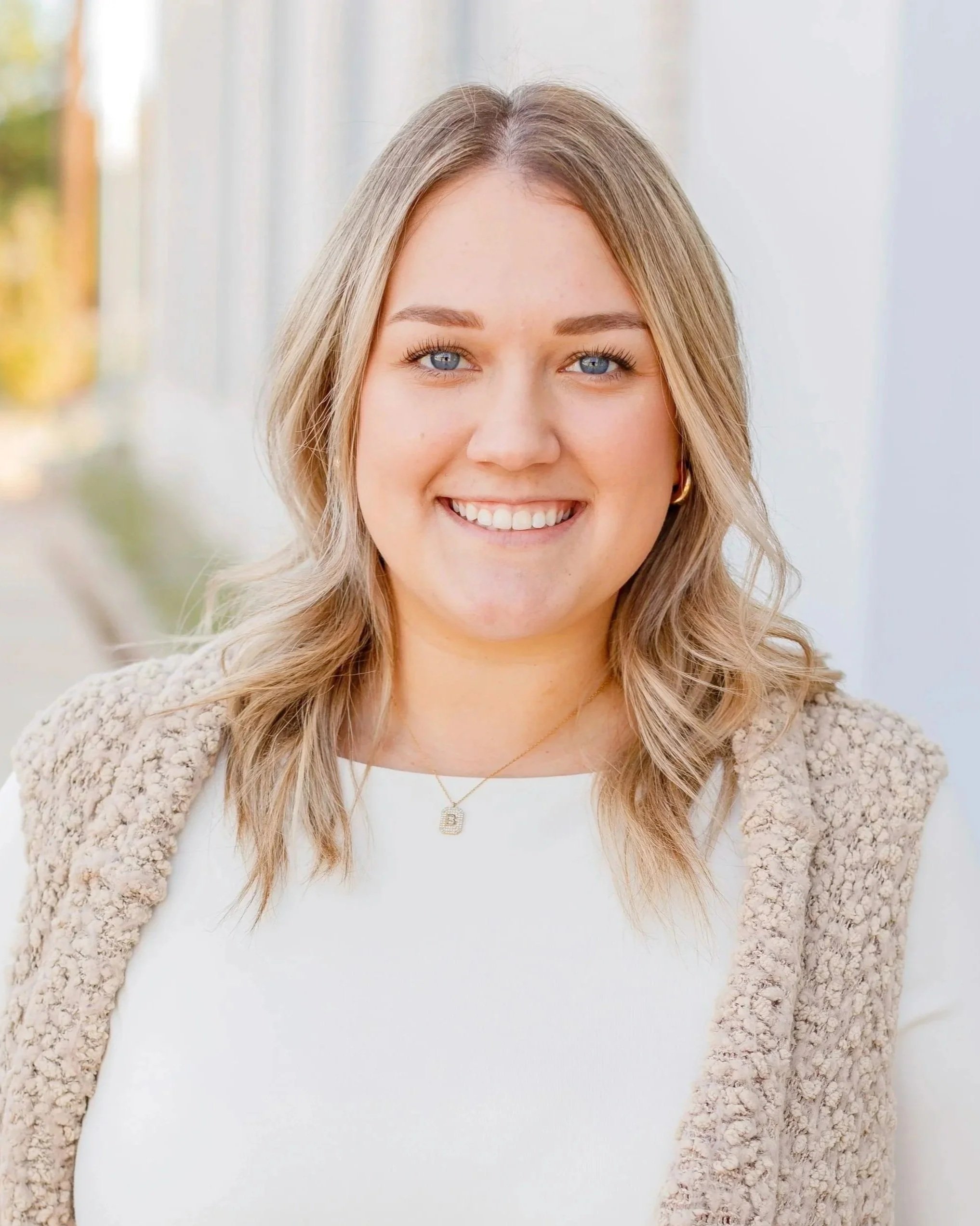 A young woman with shoulder-length blond hair and blue eyes smiling outdoors, wearing a beige textured vest over a white top, and a gold necklace with a letter 'B' pendant.