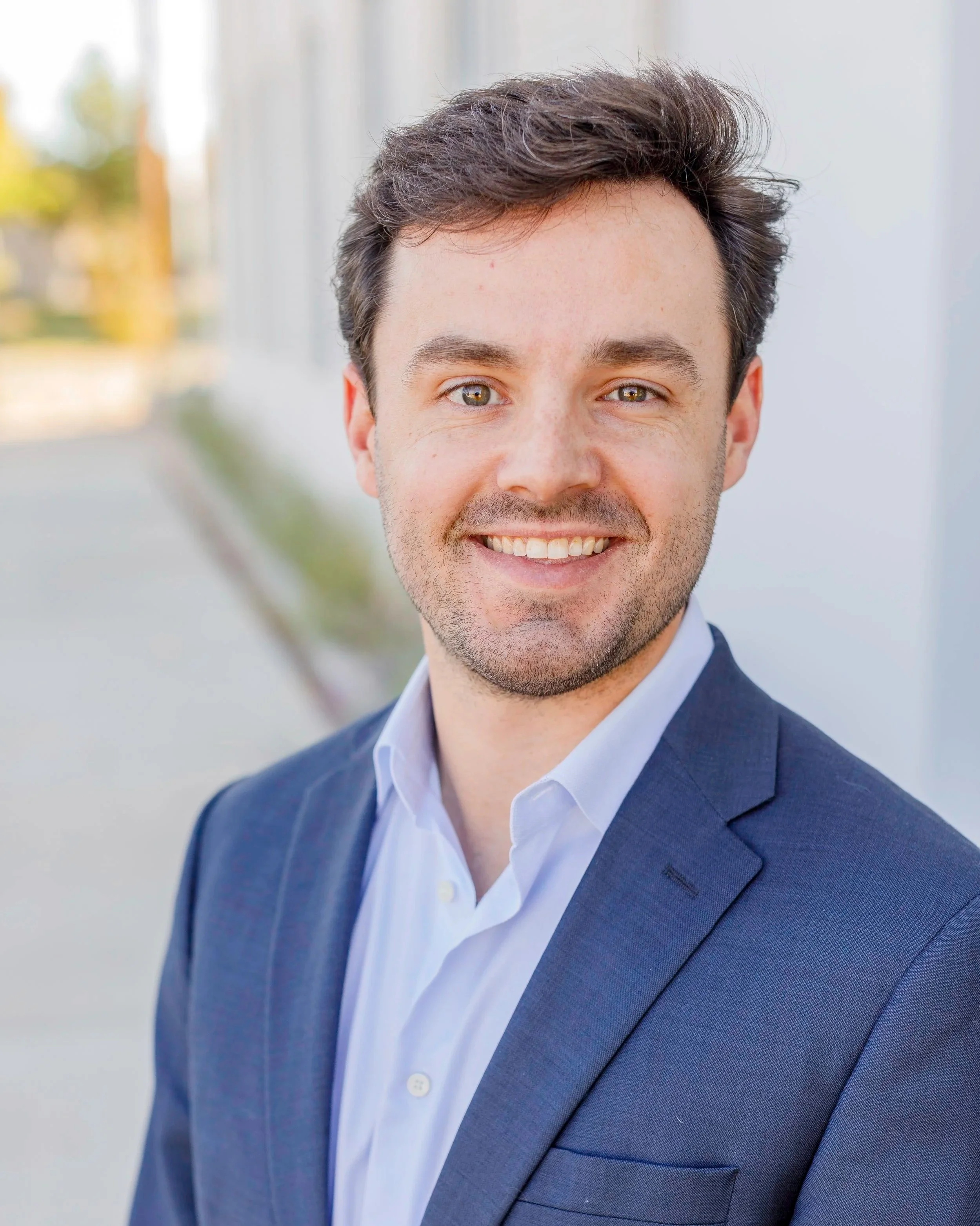 Headshot of a smiling man in a suit, outdoors with blurred background.
