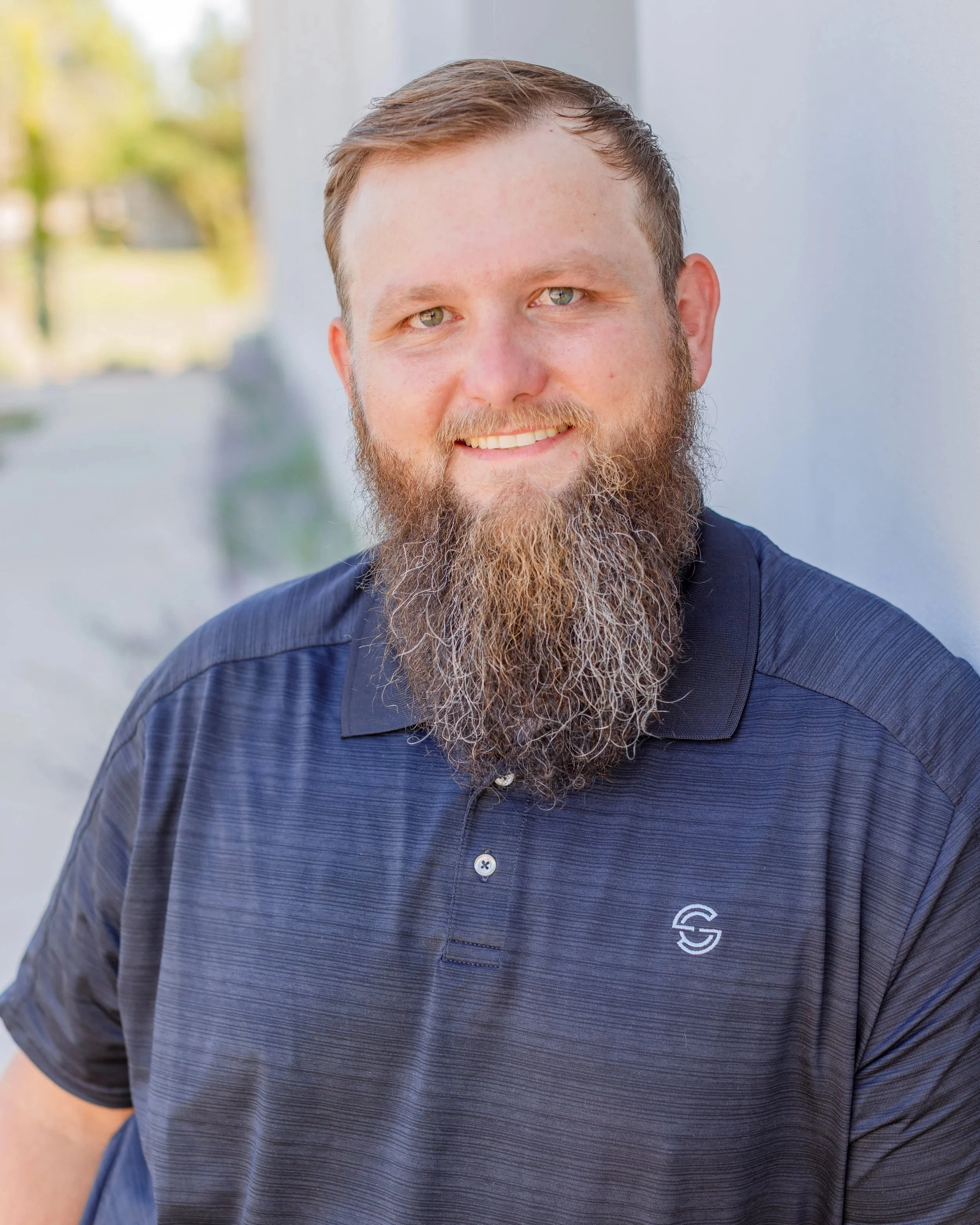 A man with a beard and short hair wearing a navy blue polo shirt, smiling outdoors on a sunny day.