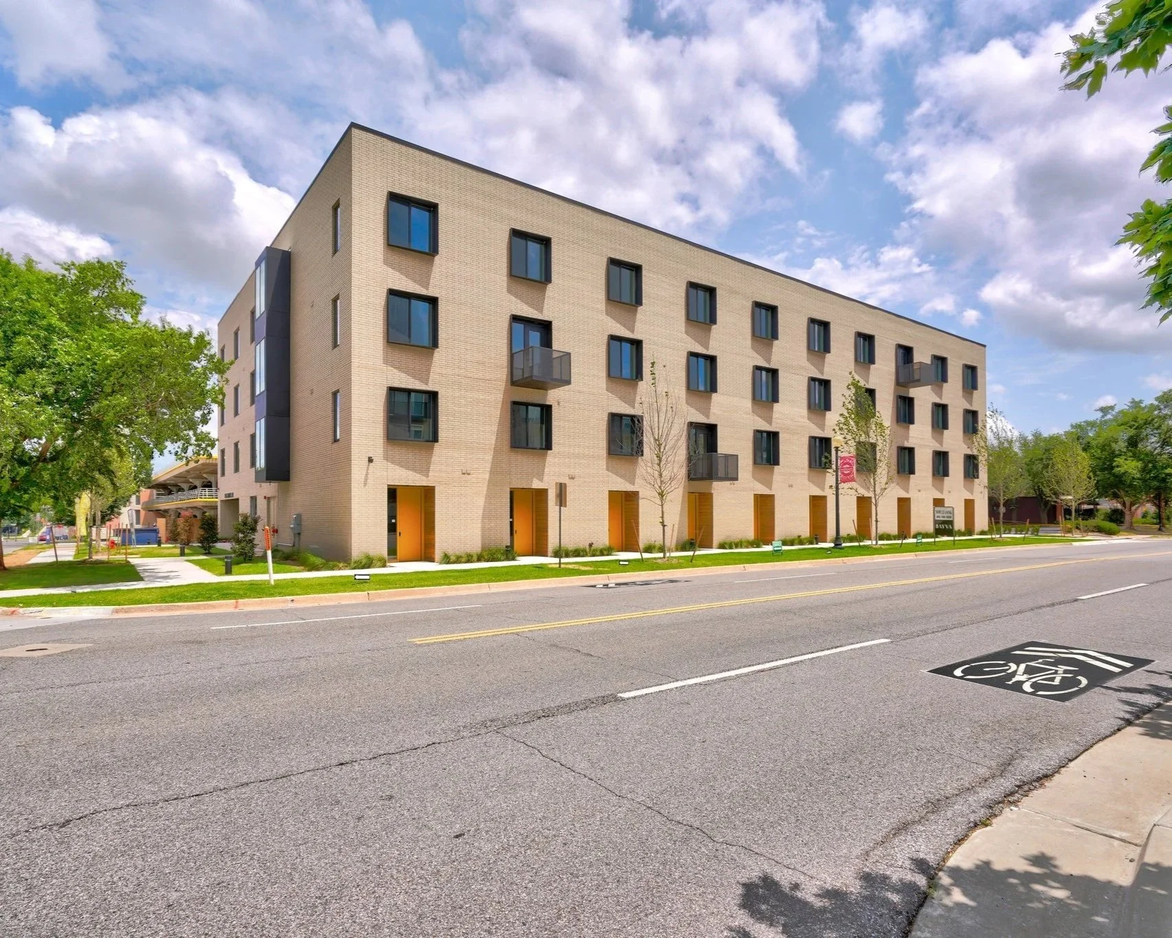A multi-story modern apartment building with beige brick exterior and black window frames, situated on a tree-lined street with a bike lane.