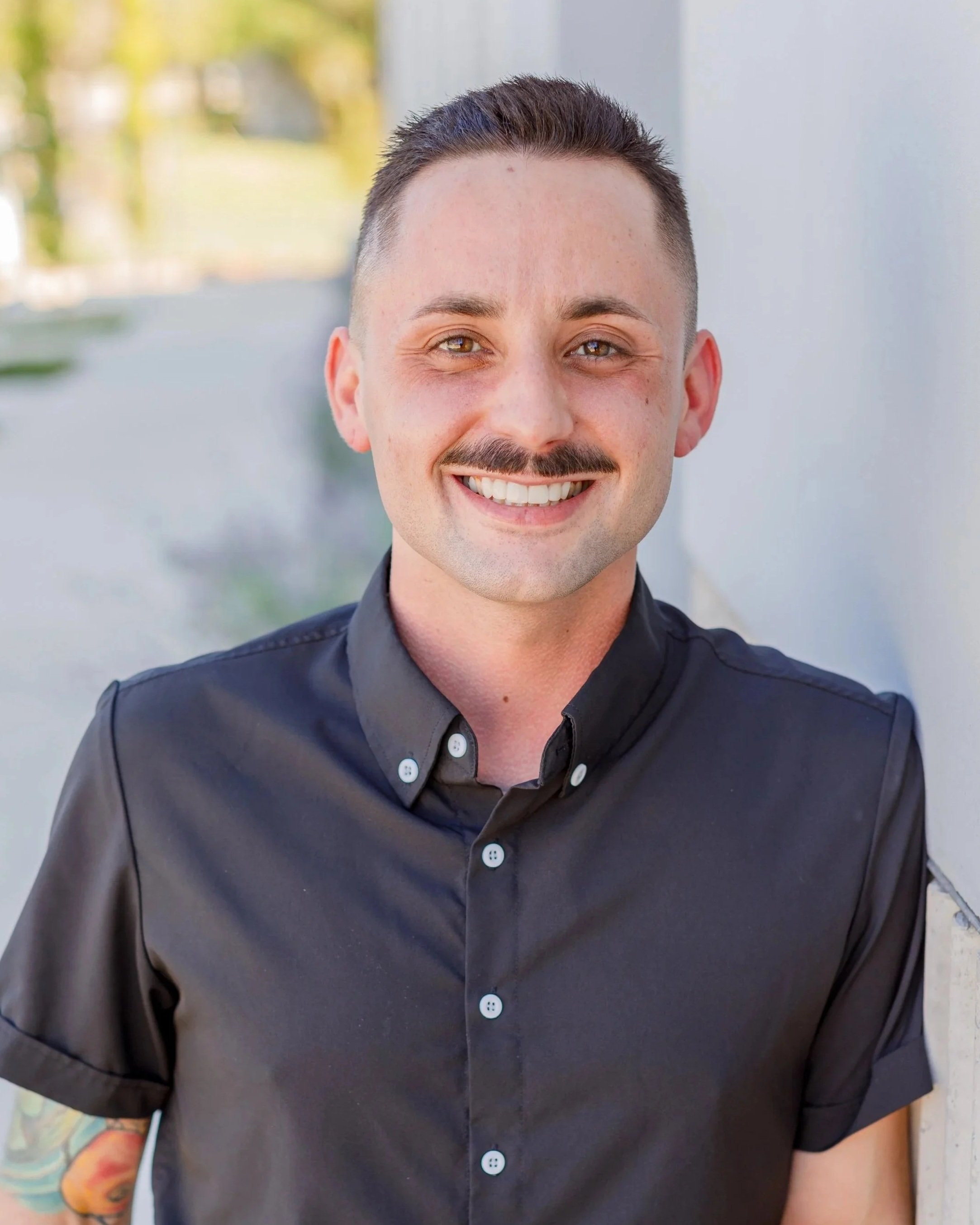 Headshot of a smiling man with short dark hair, a mustache, wearing a black button-up shirt, outdoors in bright daylight.