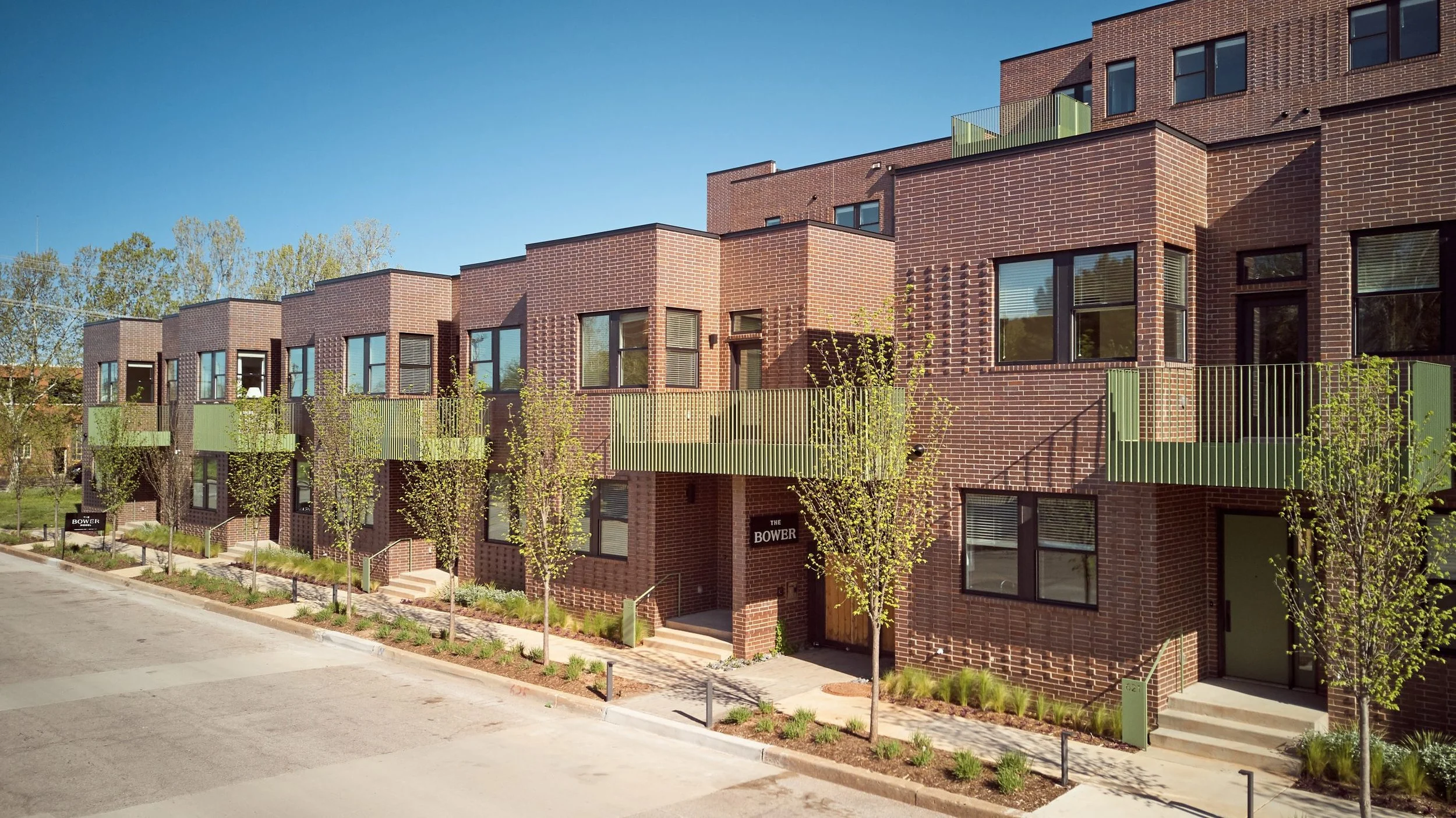 Modern brick apartment building with green balconies, trees lining the sidewalk, and a clear blue sky.