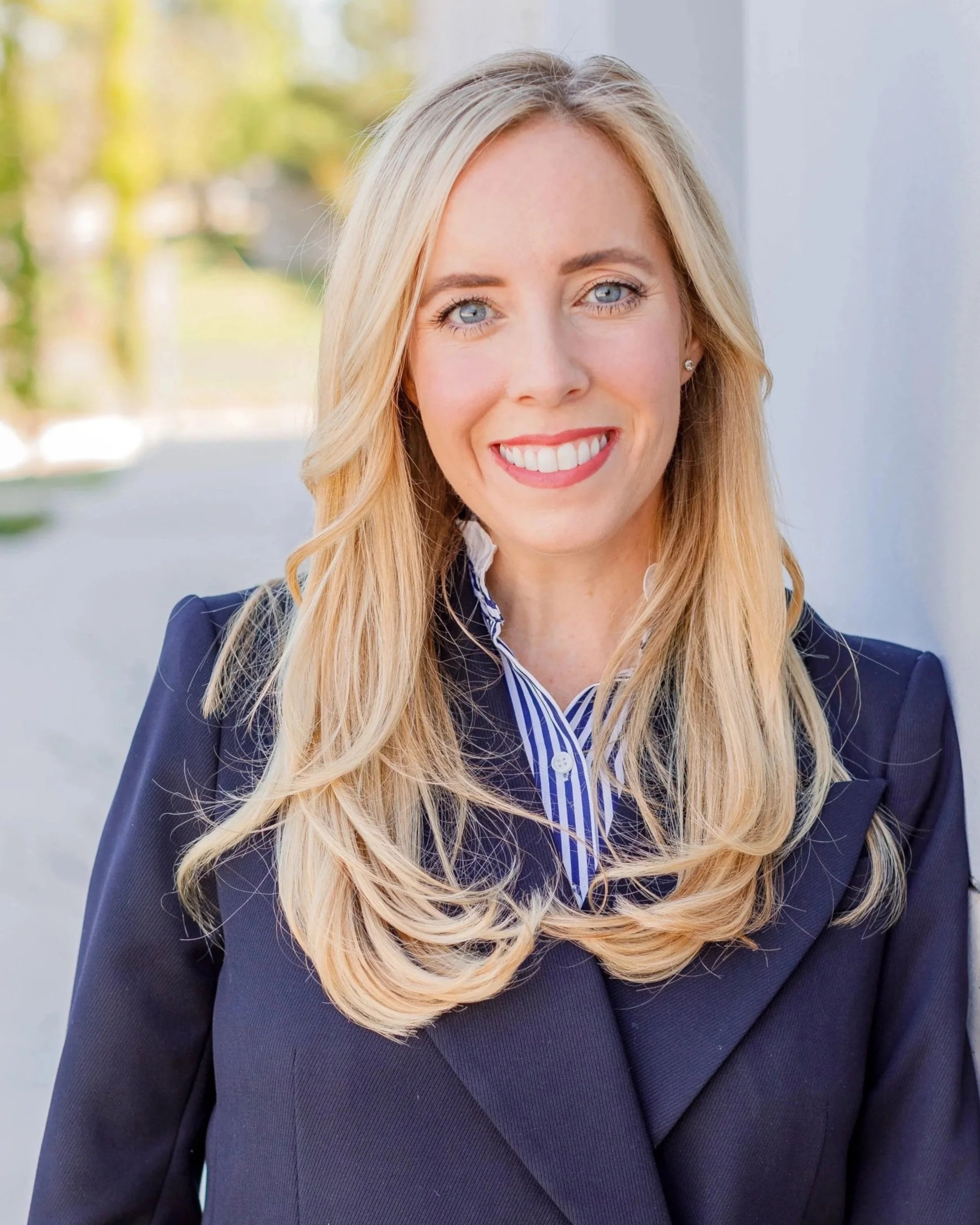 A woman with long blonde hair wearing a dark blazer and a blue and white striped shirt, smiling outdoors.