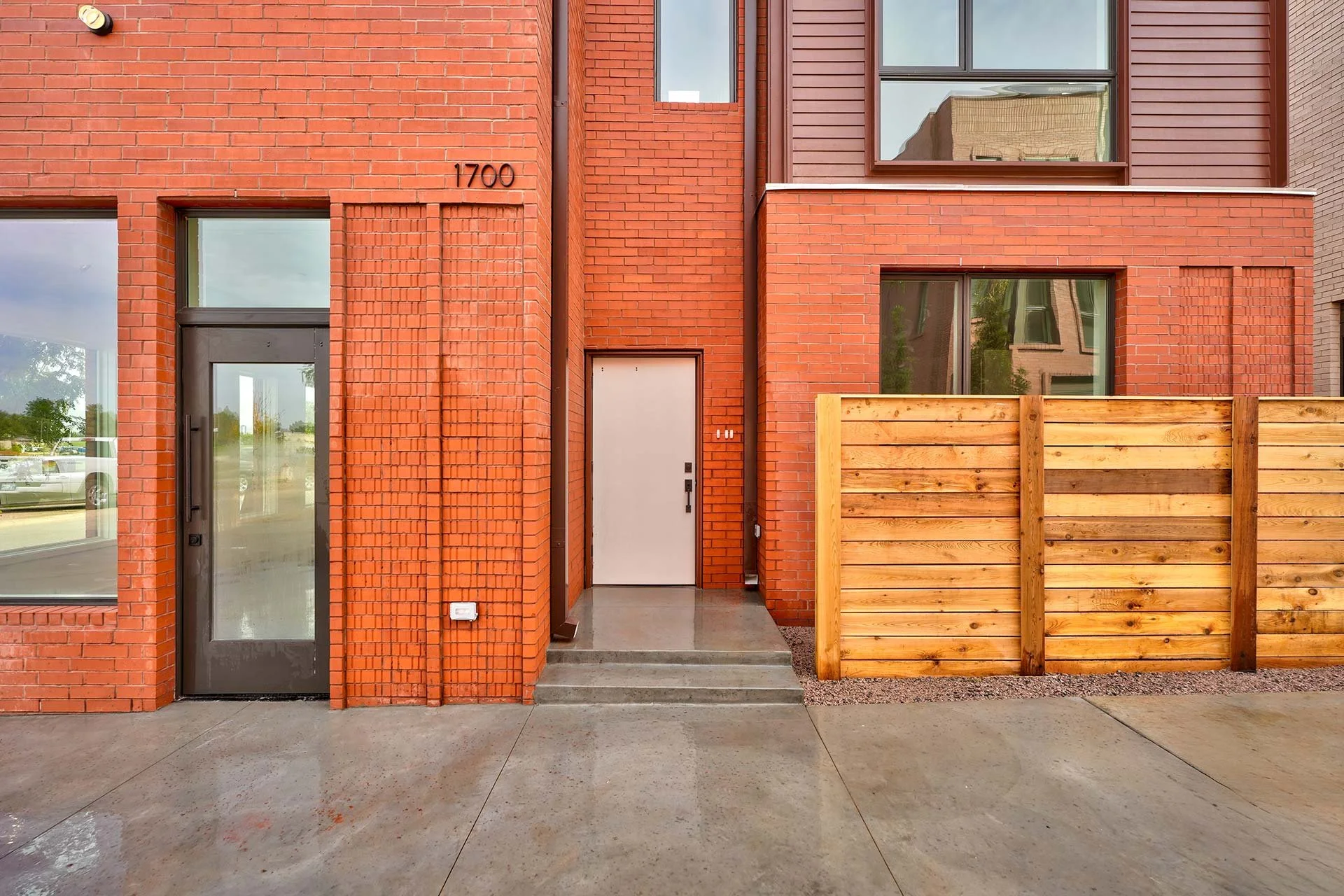Exterior view of a modern brick building with the number 1700, featuring glass doors and windows, a wooden privacy fence on the right, and concrete sidewalk.